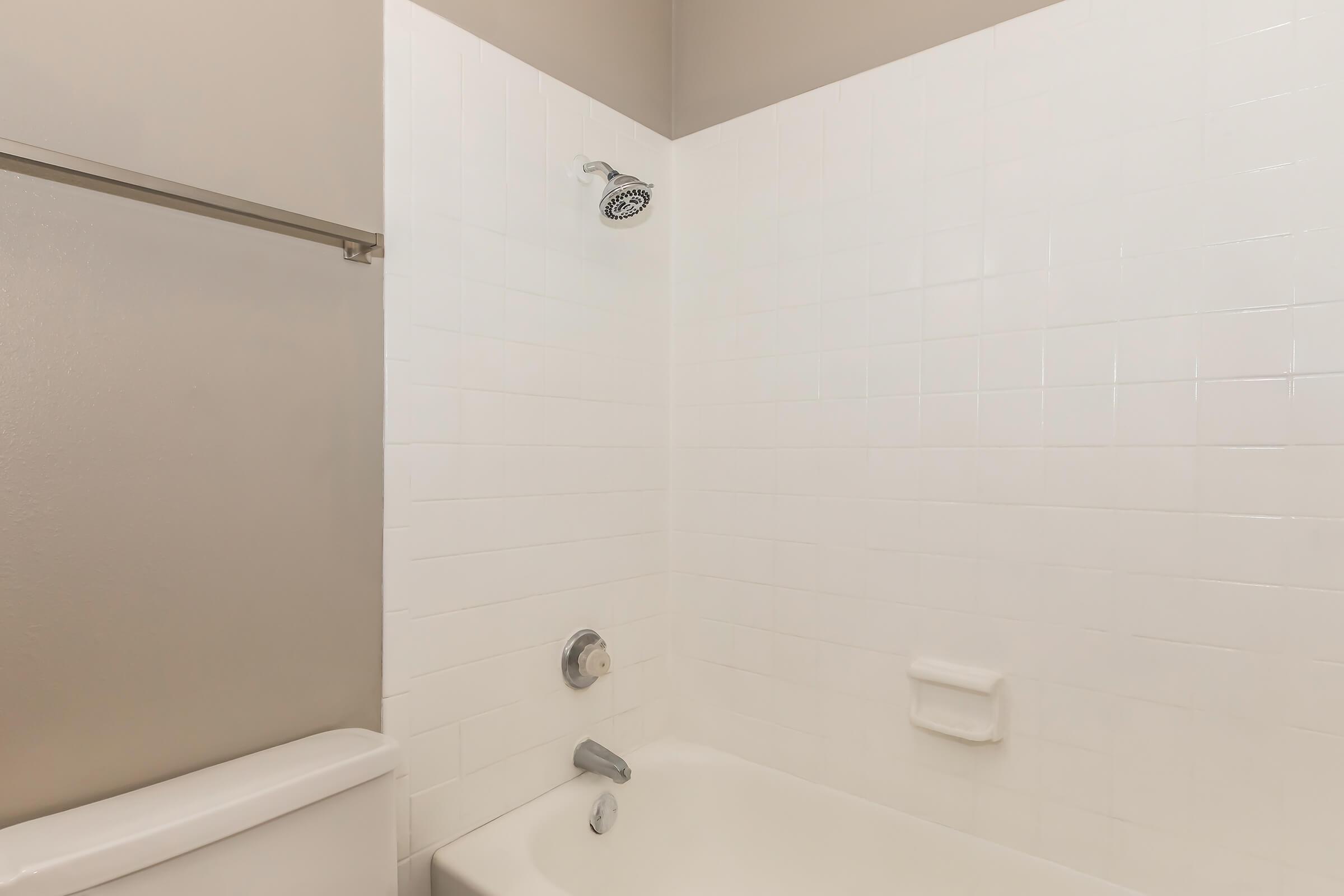 A clean bathroom featuring a white tiled shower area with a showerhead and a white bathtub. The walls have a neutral paint color, and there is a towel rack on the left. The overall design is minimalist and modern.
