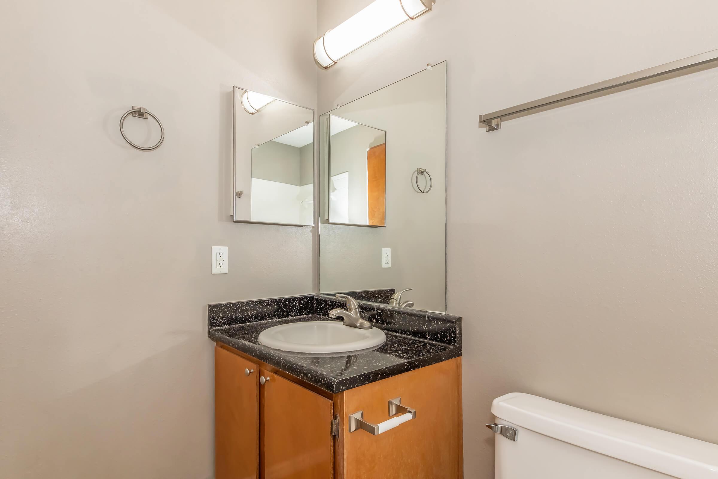 A modern bathroom featuring a stylish sink with a granite countertop, a large mirror above the sink, wooden cabinetry, a towel ring, and a white toilet. The walls are painted in a soft neutral color, and there is a light fixture above the sink, providing ample illumination.