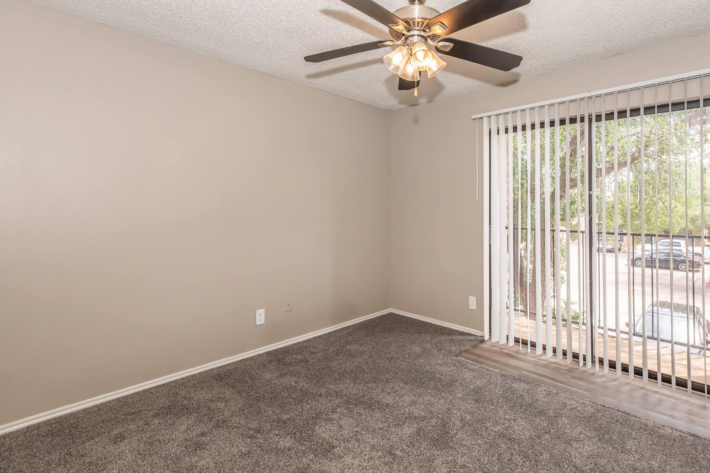 An empty room with light gray walls and carpet, featuring a ceiling fan with light. There are vertical blinds on a window that overlooks an outdoor area with trees and parked cars. The space is well-lit and has a simple, clean aesthetic.