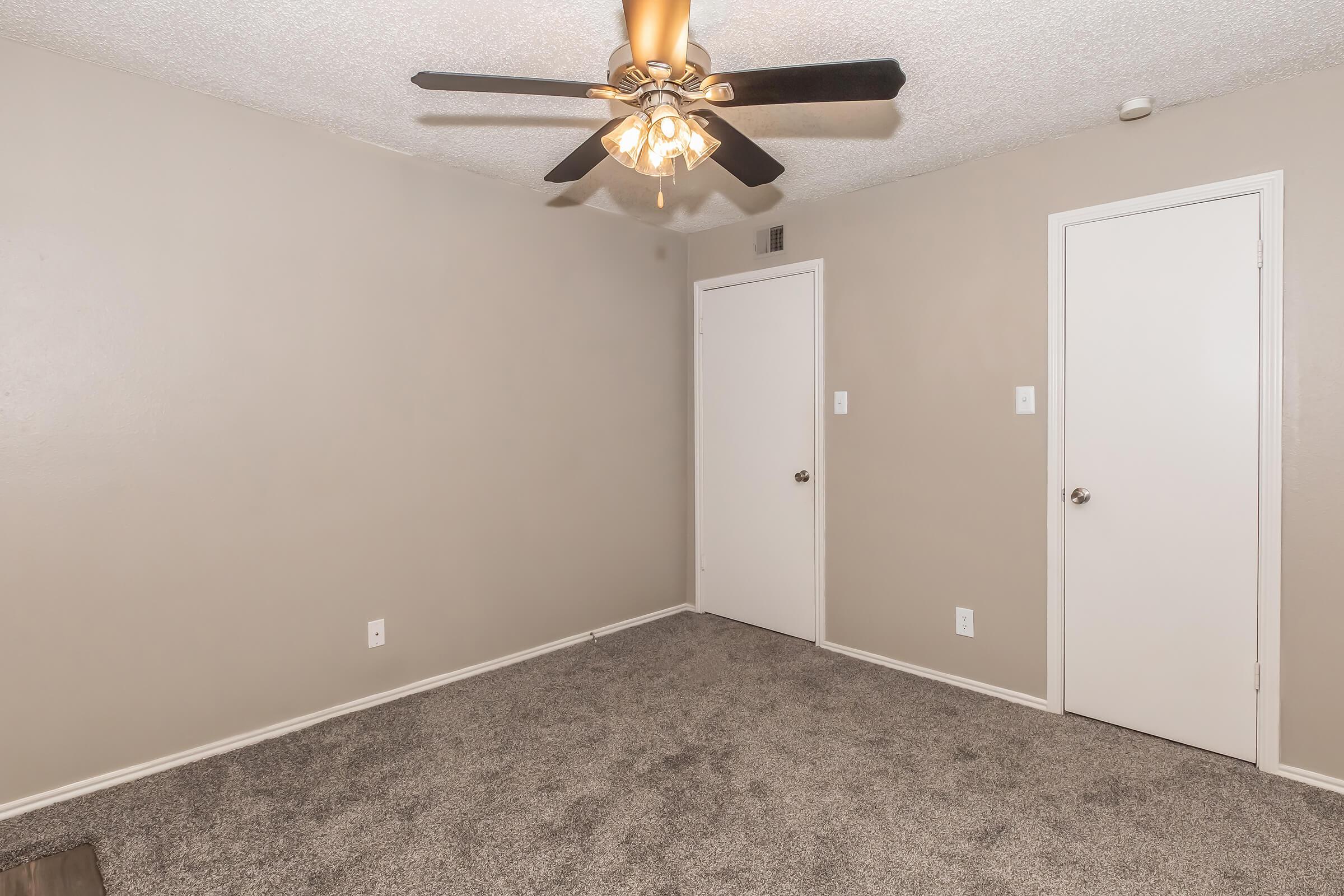 A well-lit room featuring light gray walls and plush gray carpeting. It has a ceiling fan with light fixtures and two closed doors on the right. The space appears empty and ready for furnishing, creating a neutral and inviting atmosphere.
