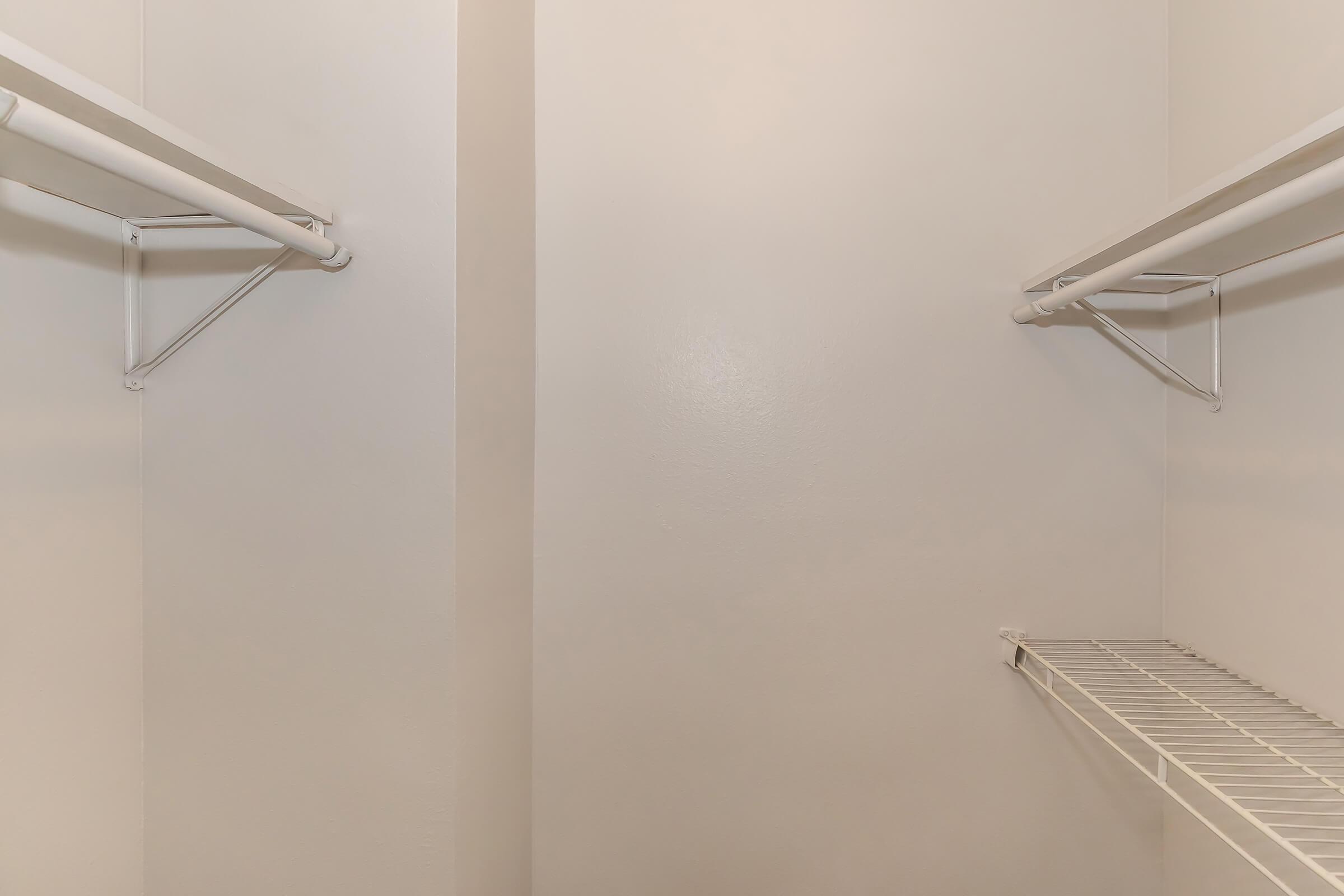 Empty closet with white walls and wire shelving. Two horizontal bars mounted on the side walls for hanging clothes, and a wire shelf on the bottom, providing storage space. The overall appearance is simple and uncluttered, highlighting the lack of items inside.