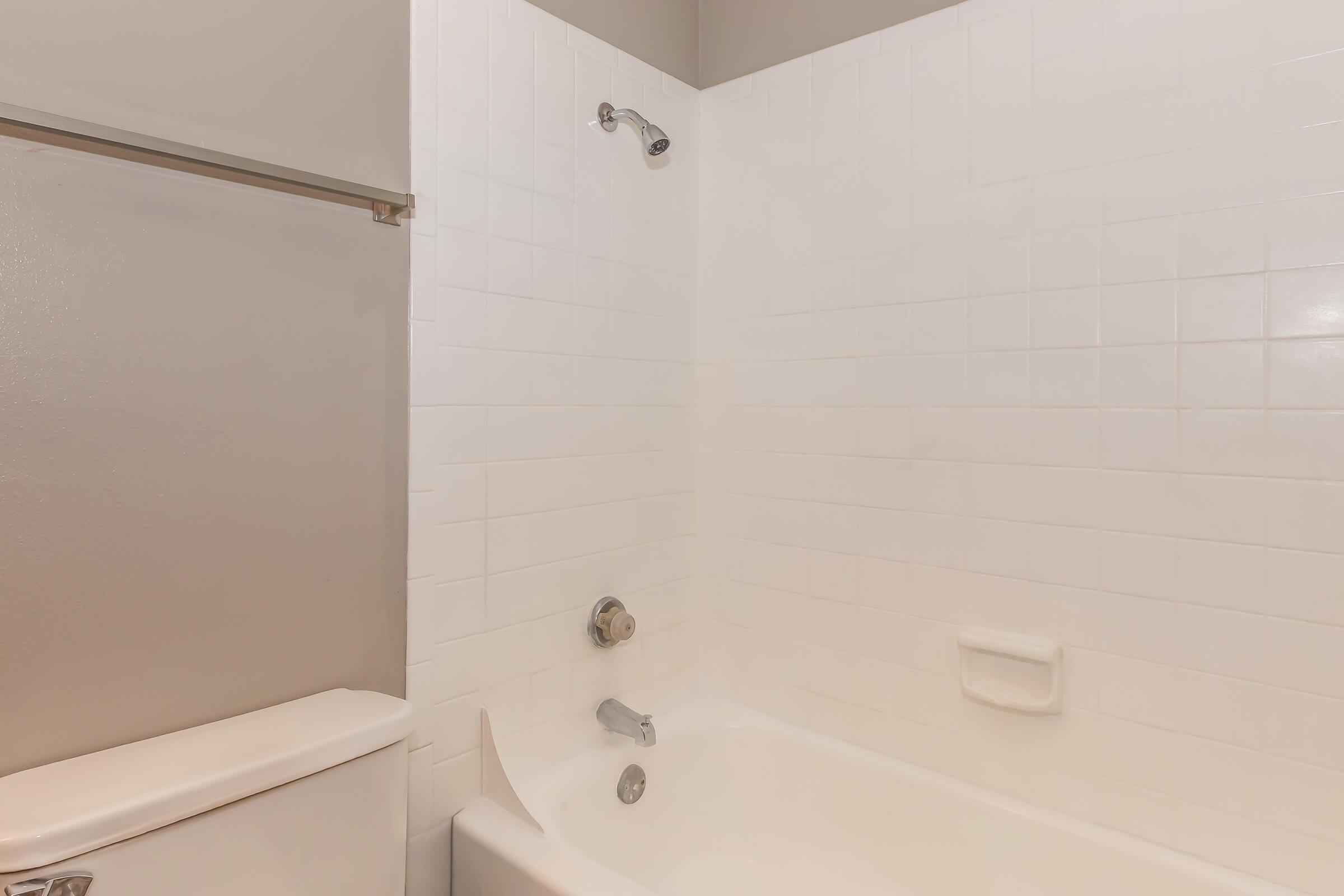 A clean, minimalist bathroom featuring a white tiled bathtub and shower with a modern faucet. A toilet is visible in the corner, and the walls are painted in a light color, enhancing the bright and spacious feel of the room. Overall, the design exhibits a contemporary style.