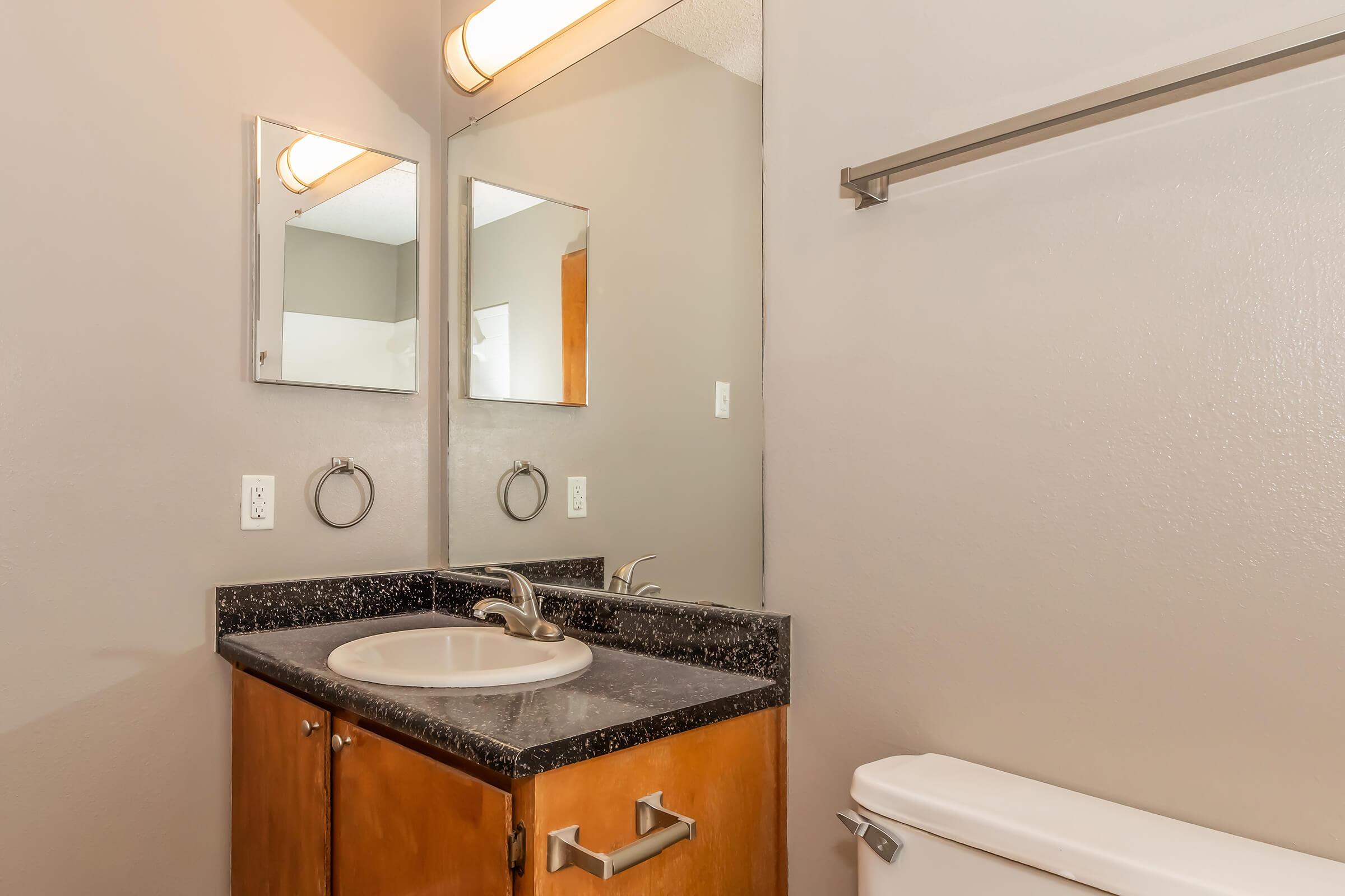 Modern bathroom featuring a double mirror above a countertop sink with a dark granite surface. The wooden vanity includes two cabinet doors and a towel ring on the wall. A toilet is positioned adjacent, with gray walls and a simple light fixture overhead.