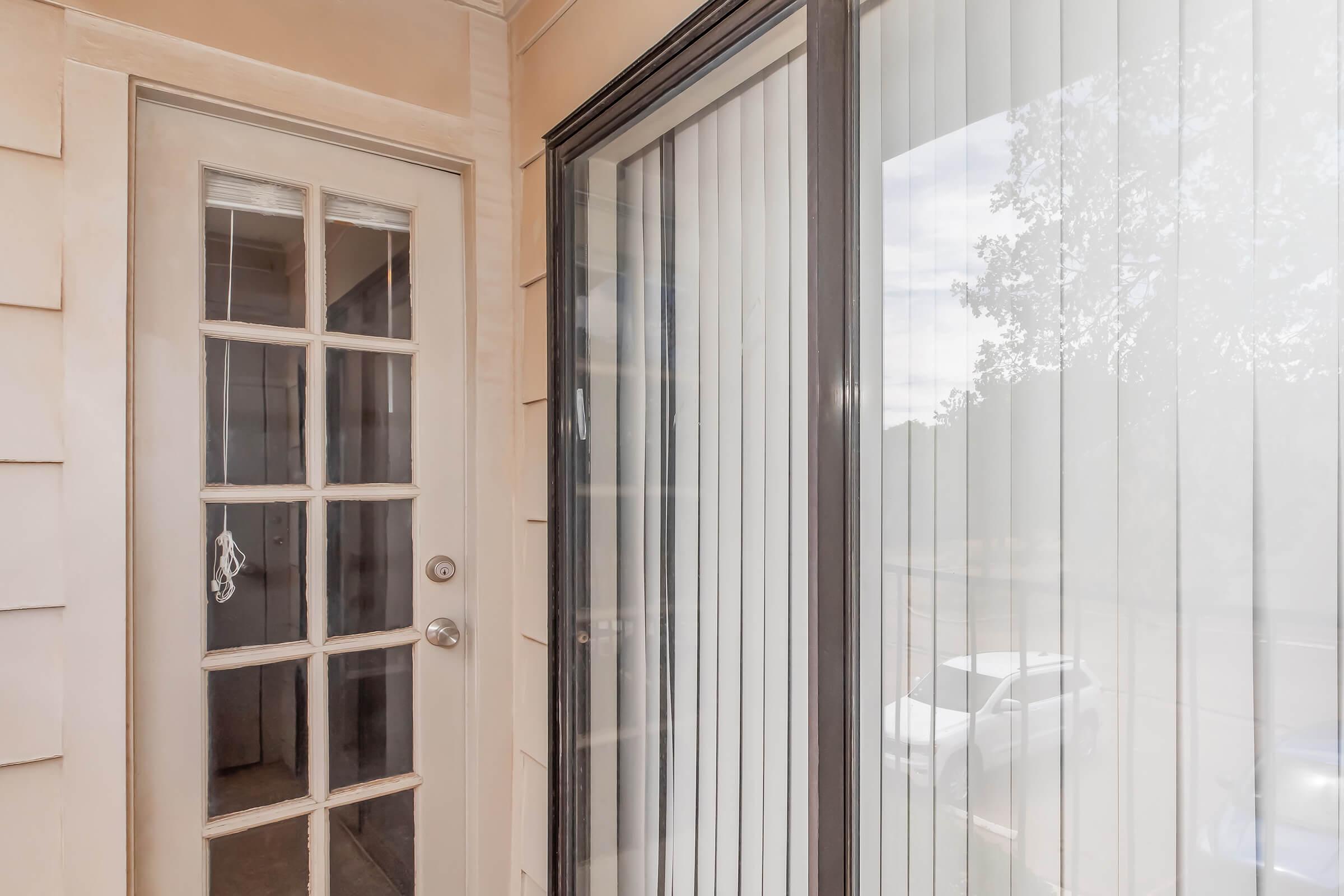 A view of a door with multiple glass panes, leading to a room behind it. Adjacent is a window with vertical blinds partially drawn, allowing some natural light to enter. The scene is framed by a neutral-colored wall, creating a clean and inviting entrance.