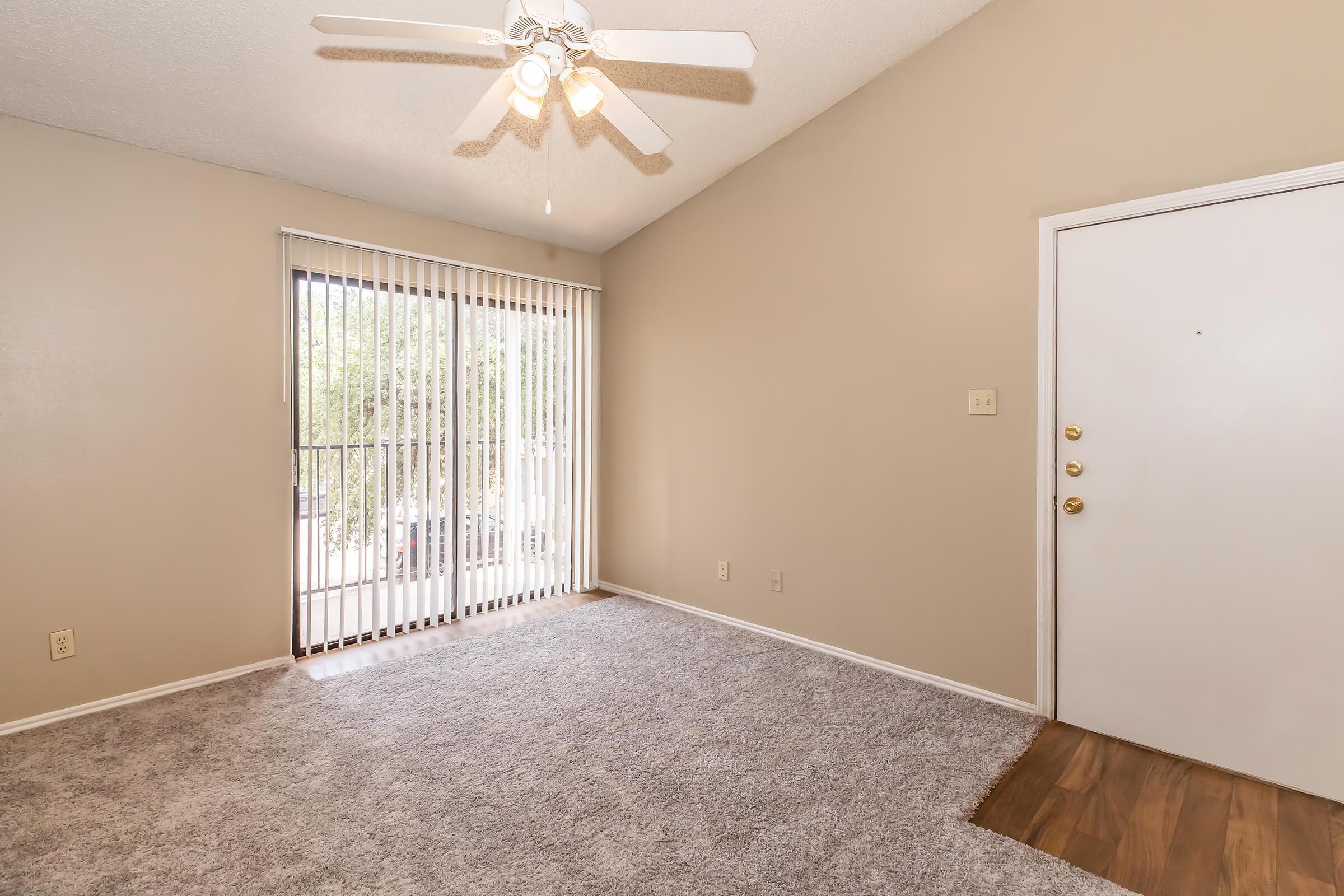 A spacious, empty living room with light beige walls and plush carpet flooring. The room features a ceiling fan and large sliding glass doors with vertical blinds allowing natural light. The entrance door is visible on the right, and the overall atmosphere is bright and airy.