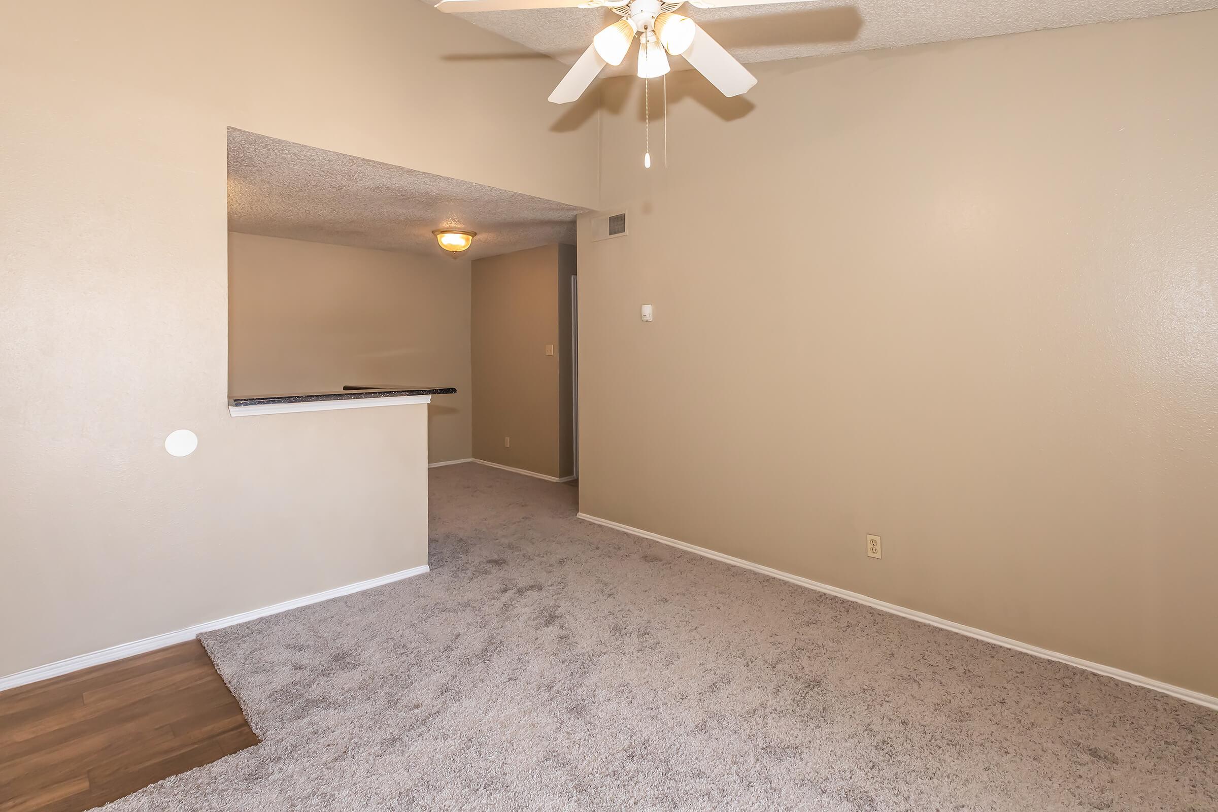 A well-lit room featuring beige walls and carpet flooring. The room includes a ceiling fan and a portion of a wall that appears to have a bar or counter opening to another space. There is a small area visible with wood flooring and no furniture.