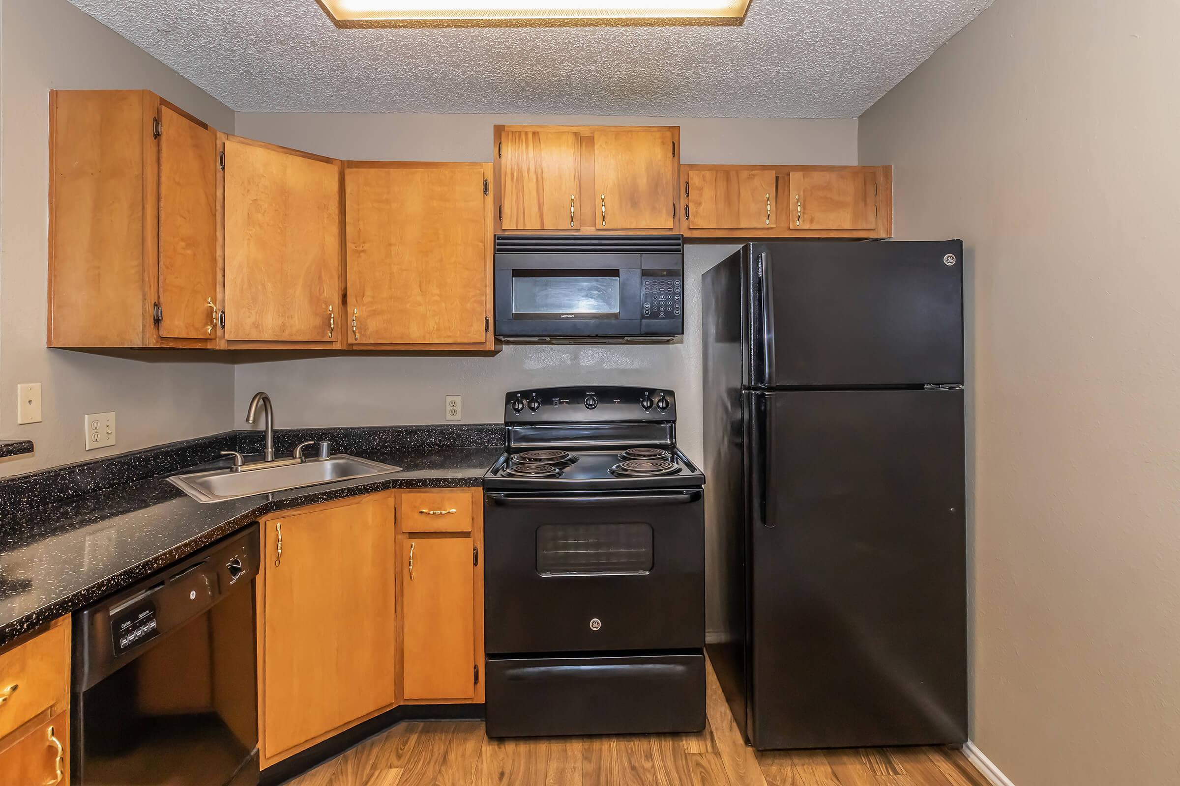 A modern kitchen featuring wooden cabinets, a black stove and microwave, a black refrigerator, and a sink. The countertops are dark granite, and the flooring appears to be wood laminate. The walls are painted a light neutral color, creating a warm and inviting atmosphere.