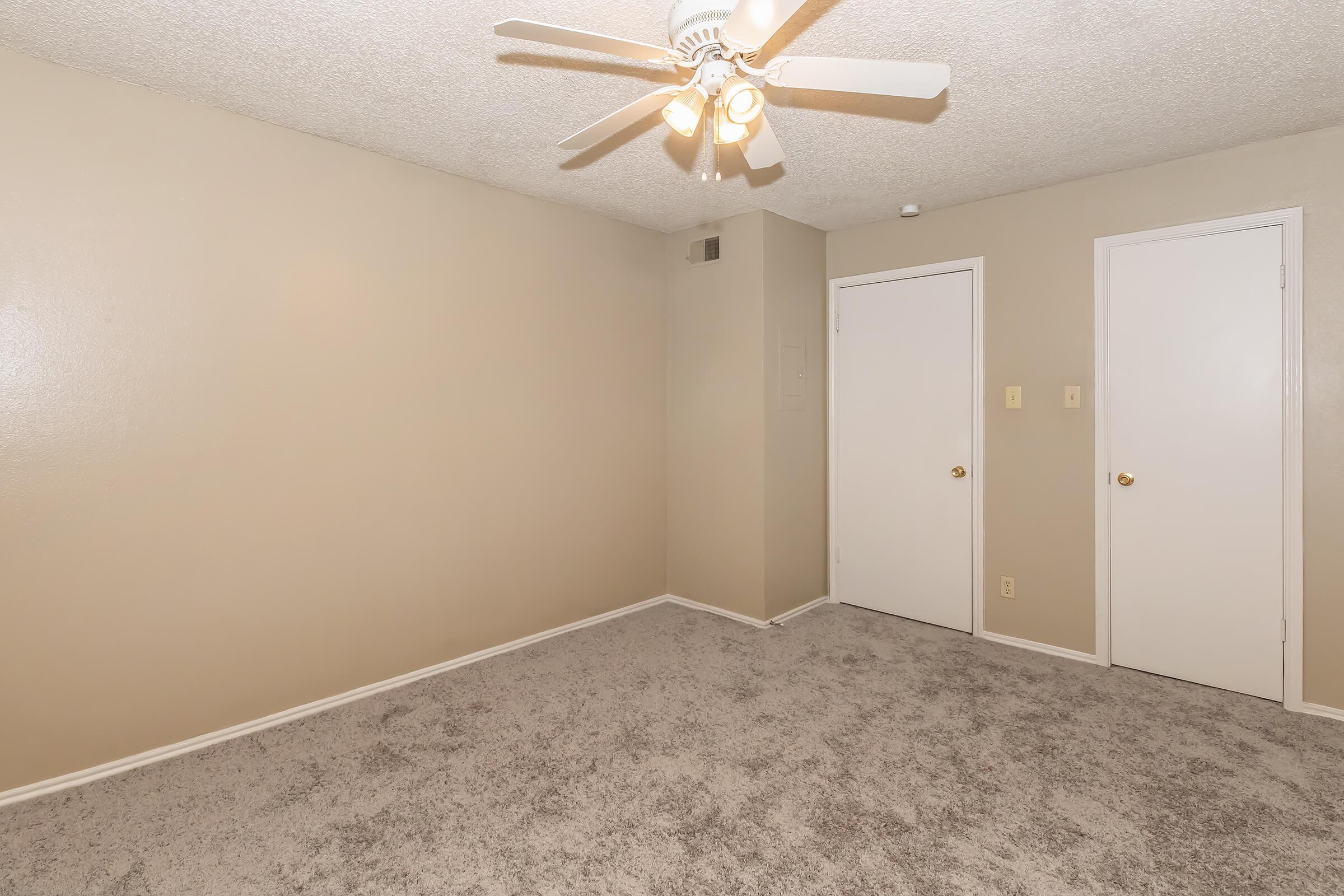 A spacious, empty room featuring light beige walls and a textured ceiling. The floor is covered in soft gray carpet. There is a ceiling fan with four light bulbs, and two closed doors on the right side of the image. The overall atmosphere is bright and airy, ideal for various uses.