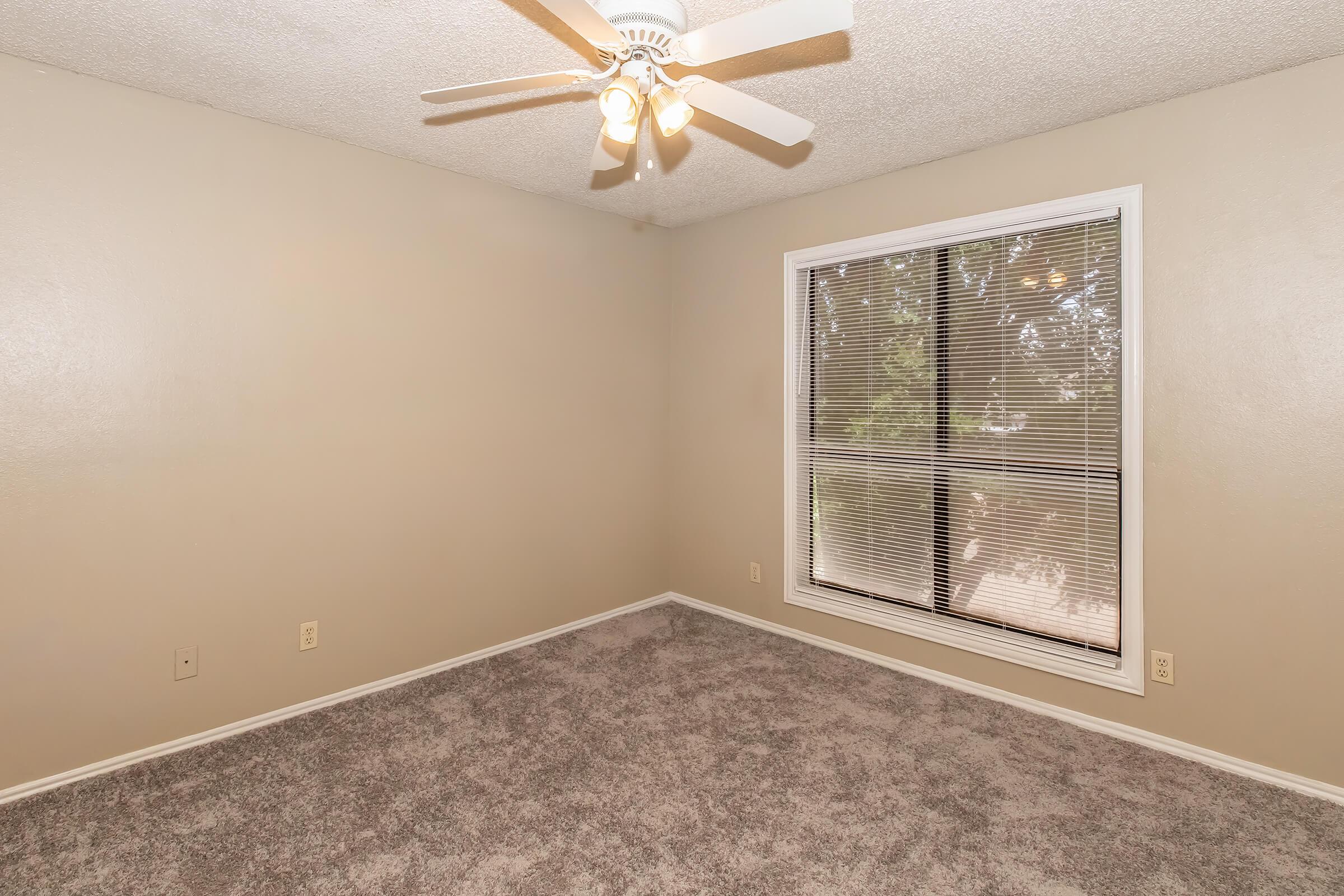 A spacious empty room with light beige walls and a ceiling fan. The floor is covered in plush gray carpet. A large window with blinds allows natural light to enter, providing a view of the outdoors. The room is unfurnished, creating an open and inviting atmosphere.
