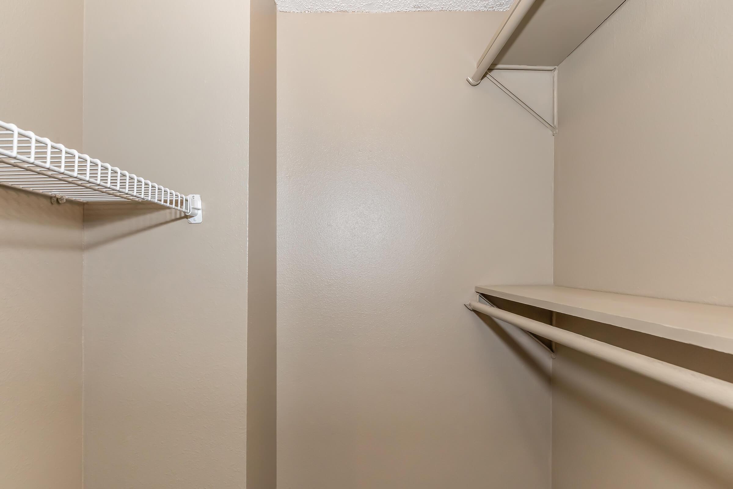Empty closet with beige walls. The space features a wire shelf attached to one wall and a flat shelf on the opposite side. The floor is clear, and there are no items or clothing present, creating a minimalist, organized appearance.