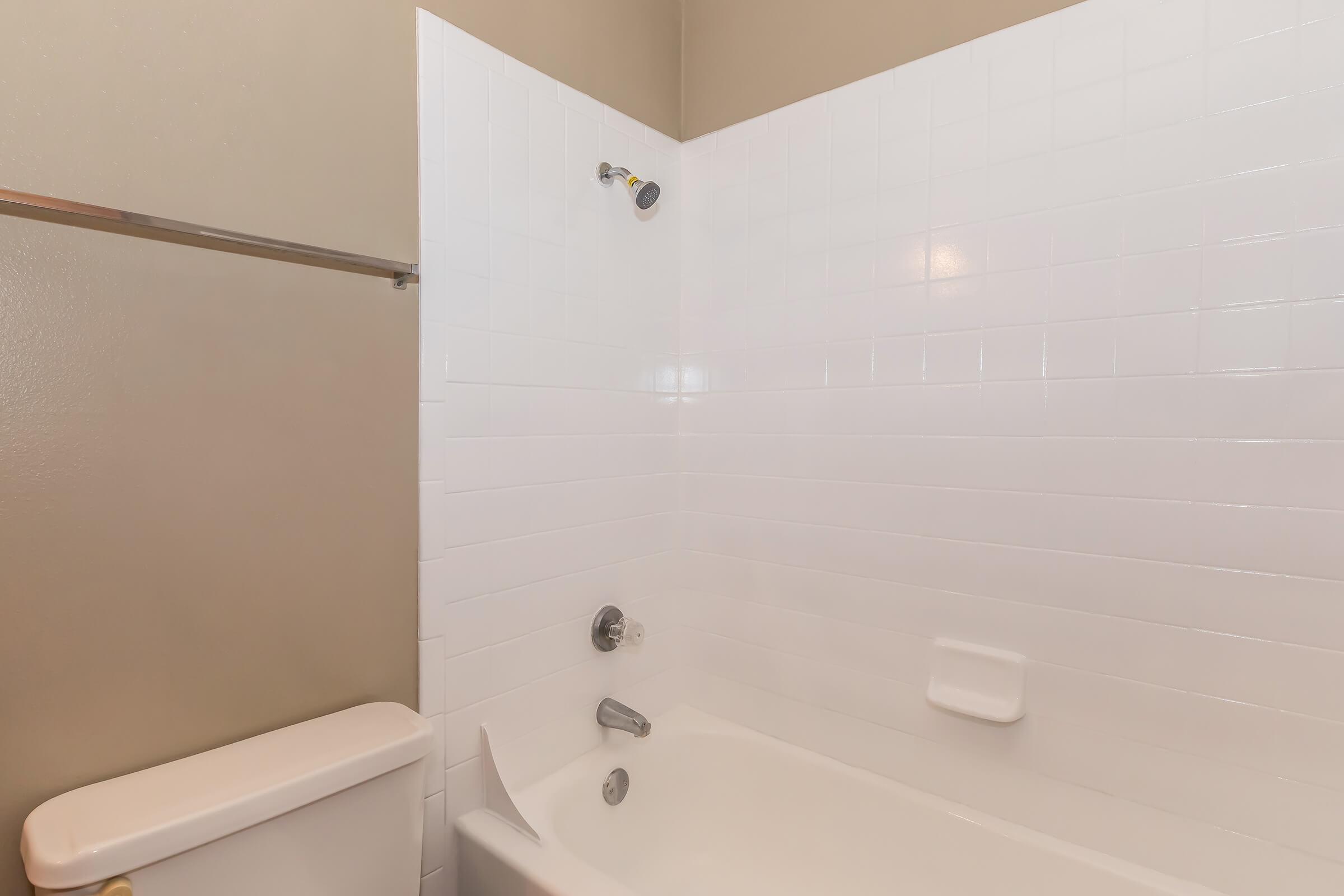 A clean, white-tiled bathroom featuring a bathtub with a showerhead above, a wall-mounted faucet, and a toilet nearby. The walls are painted a light color, creating a bright and spacious atmosphere. The space is uncluttered, emphasizing simplicity and functionality.