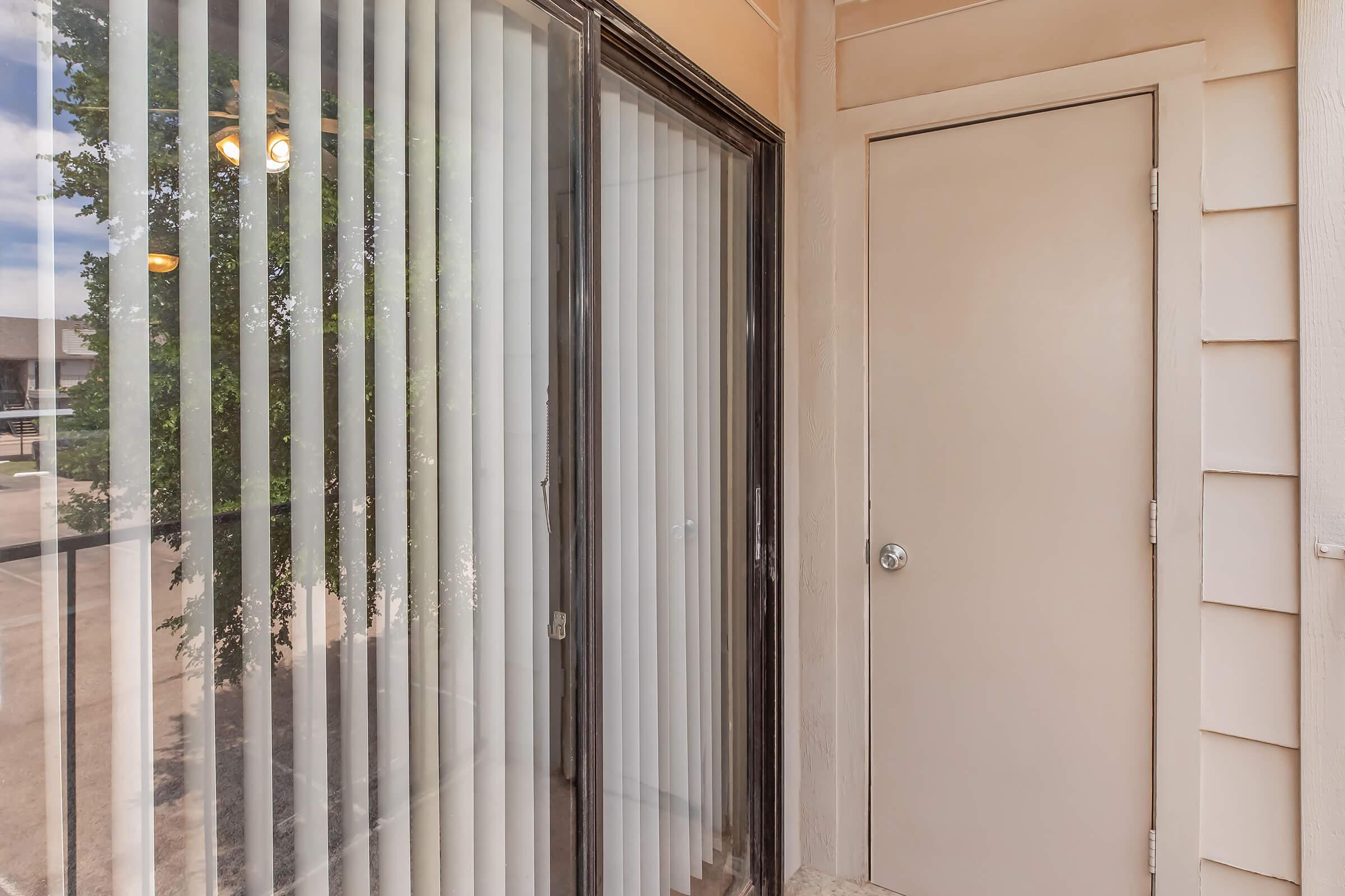 A view of an exterior balcony or porch showcasing sliding glass doors with vertical blinds, leading to a room. To the side, there is a closed door that might connect to another area. The surrounding architecture features light-colored walls and a clear blue sky is visible through the blinds.
