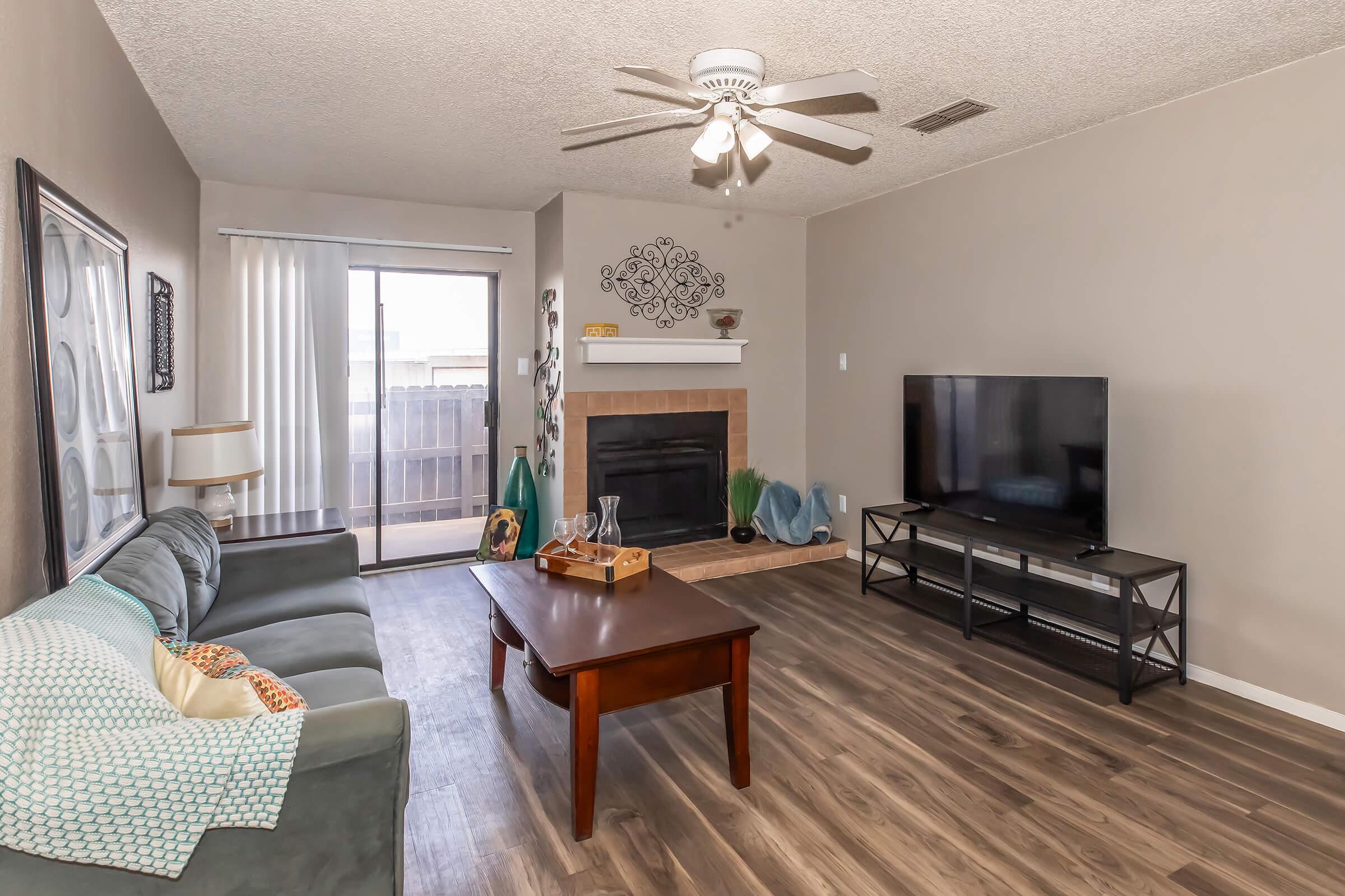 A cozy living room featuring a light blue couch with decorative pillows, a wooden coffee table with a tray, a modern flat-screen TV on a black stand, and a fireplace with decorative items. Large sliding glass doors lead to a balcony, and the room has neutral-colored walls and wooden flooring.
