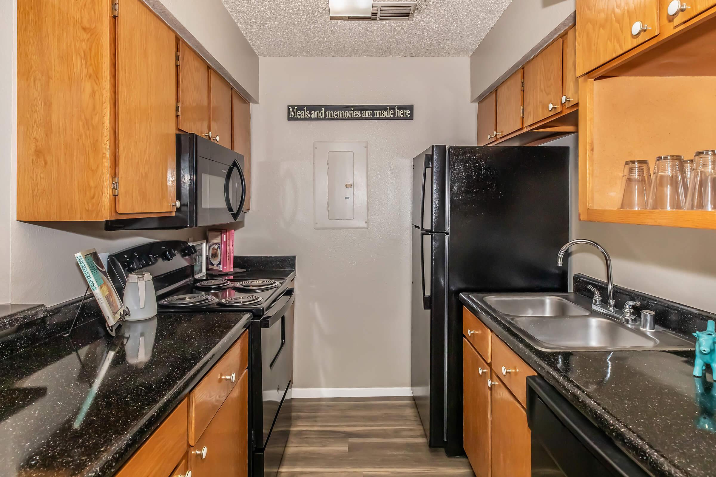 A modern kitchen featuring wooden cabinets, black appliances including a microwave, oven, and refrigerator. The countertops are dark, with a double sink visible. There is decor on the wall above the sink that reads, "Meals and memories are made here." The floor has a wood-like appearance.