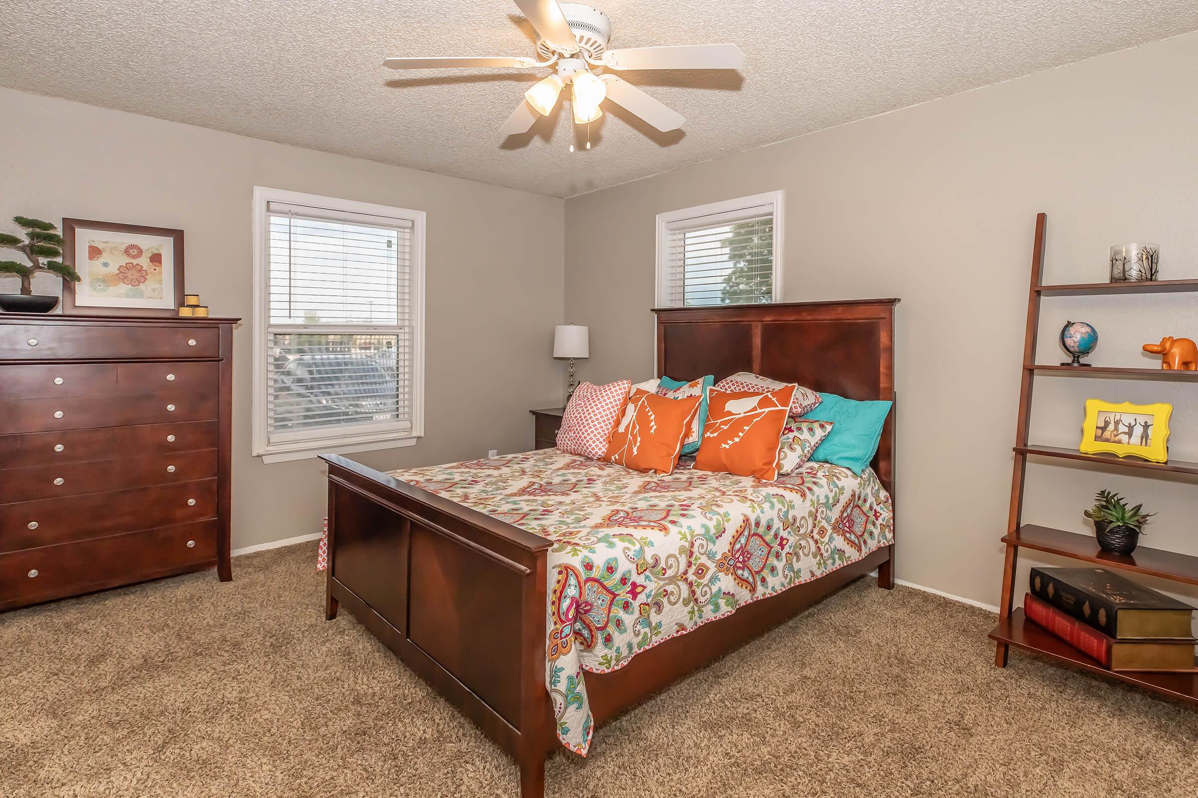 A cozy bedroom featuring a wooden bed with a colorful floral comforter and orange decorative pillows. There’s a brown dresser, a bookshelf with decorative items, and a lamp by the bed. The room has light gray walls and two windows with blinds, providing a warm and inviting atmosphere.