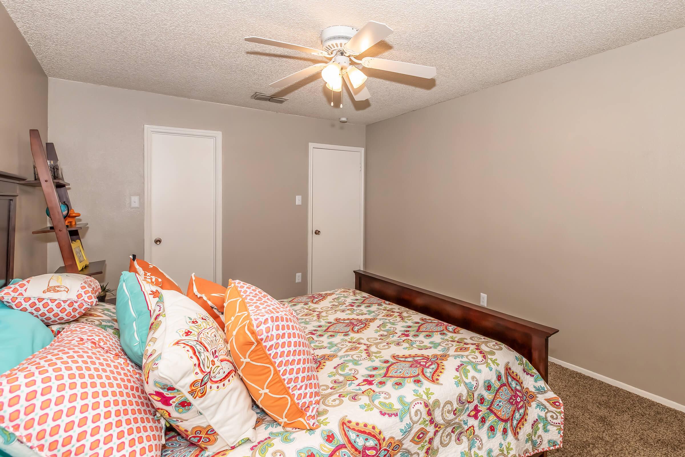 A cozy bedroom featuring a large bed with a colorful patterned duvet and an array of decorative pillows. The walls are painted a neutral color, and there are two closed doors and a wooden bench at the foot of the bed. A ceiling fan is installed, adding a touch of comfort to the space. Soft carpet covers the floor.