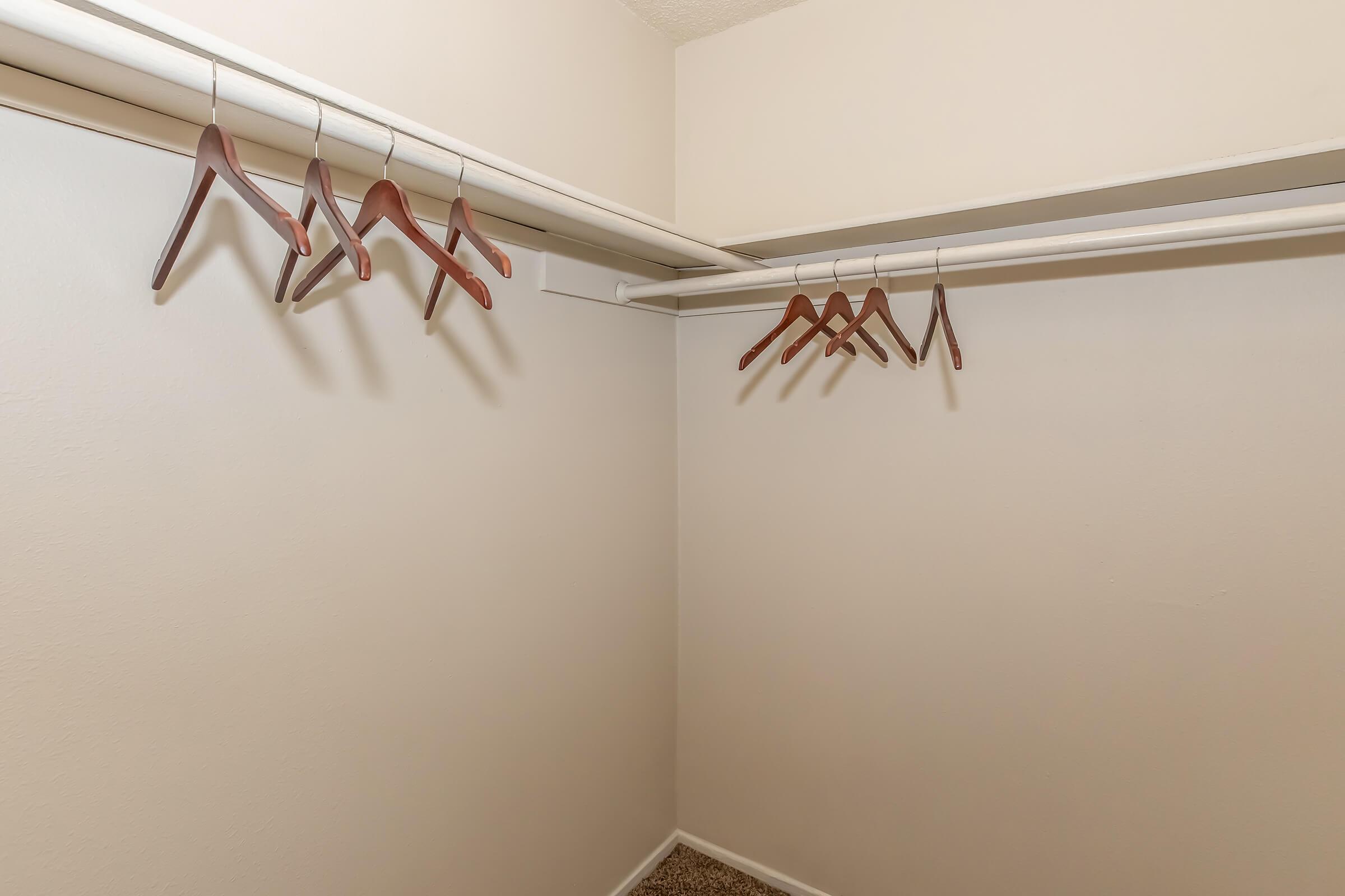 A spacious, empty closet with wooden hangers hanging from a top bar. The walls are bare and painted a light color, while the floor is carpeted. The overall look is minimalist and organized, suggesting a lack of clothing or items currently stored in the closet.