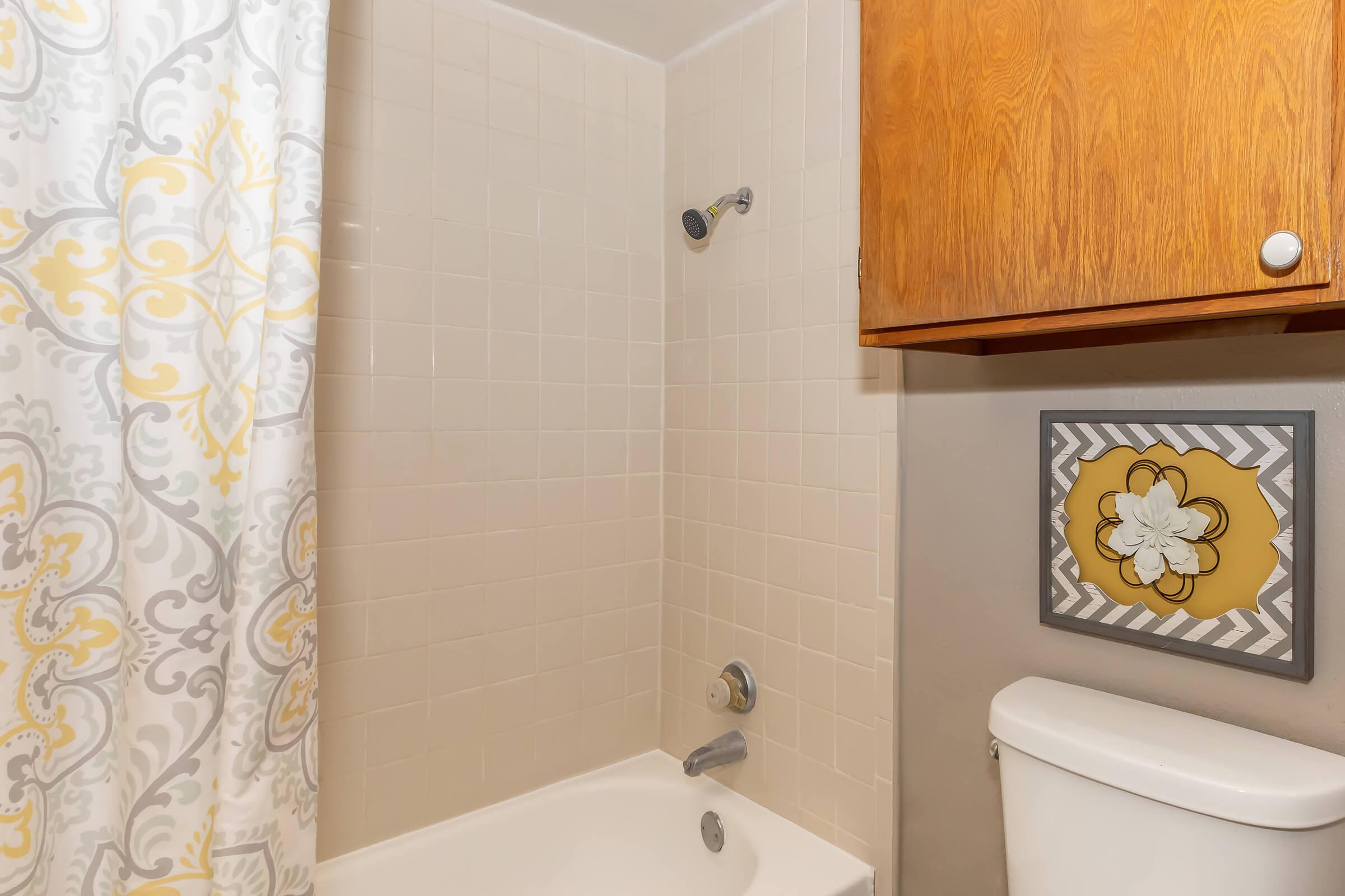 A bathroom with a bathtub and a shower. The wall features a patterned shower curtain in gray and yellow. There's a wall cabinet made of wood above the toilet, which is white. A framed decorative piece with a flower design is mounted on the wall beside the bathtub.