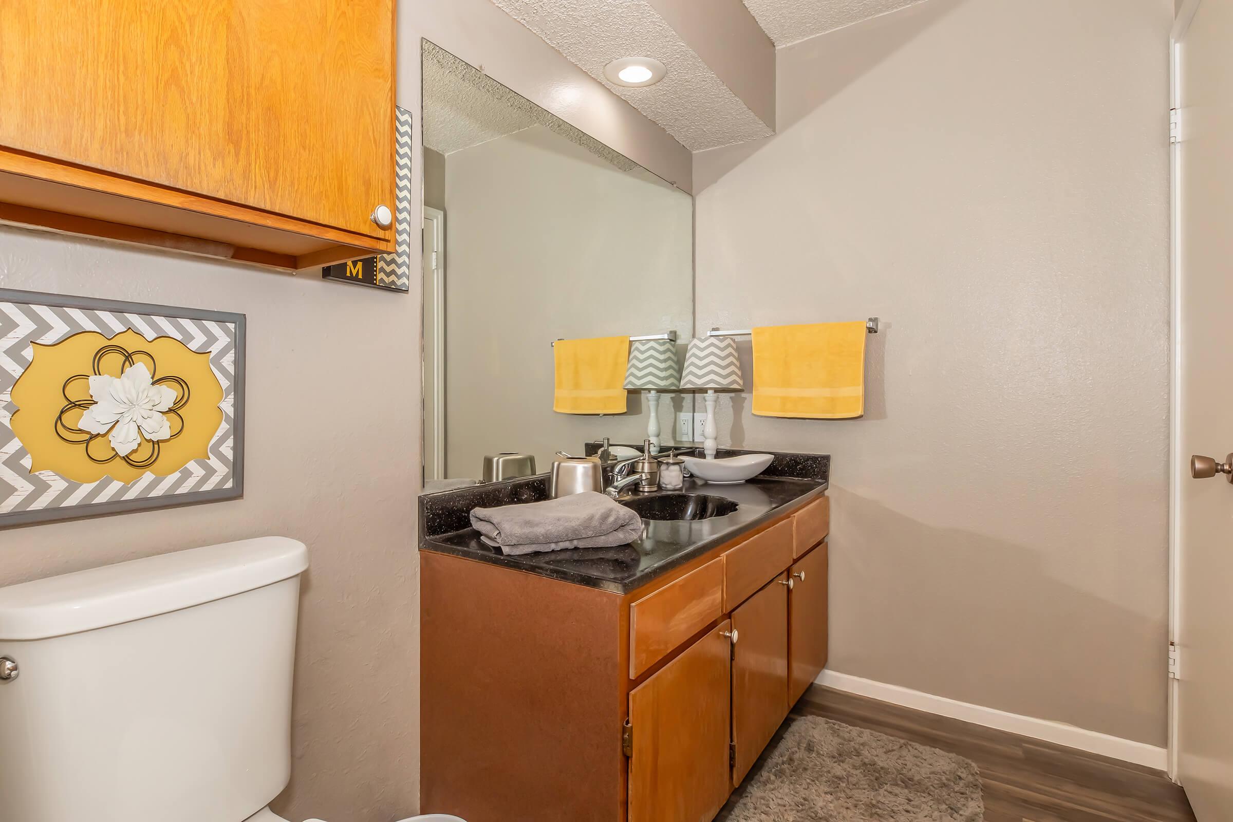 A modern bathroom featuring a wooden cabinet with yellow towels hanging on it, a rectangular mirror above a black countertop with a sink, and a toilet to the left. The wall is painted in light gray, and there's a decorative wall art piece with a flower design. A gray rug is placed on the floor.
