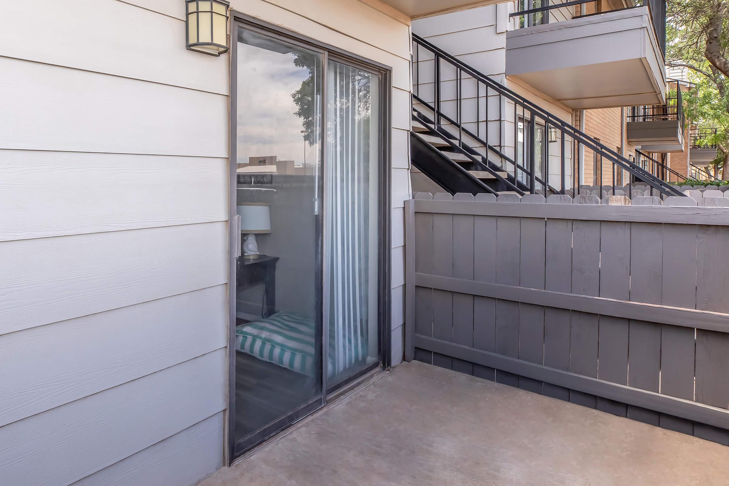 A small private patio area with a sliding glass door leading into an apartment. The patio is enclosed by a wooden fence, and there’s a glimpse of a lamp and striped bedding visible through the glass. In the background, there are steps leading to a second level of the building.