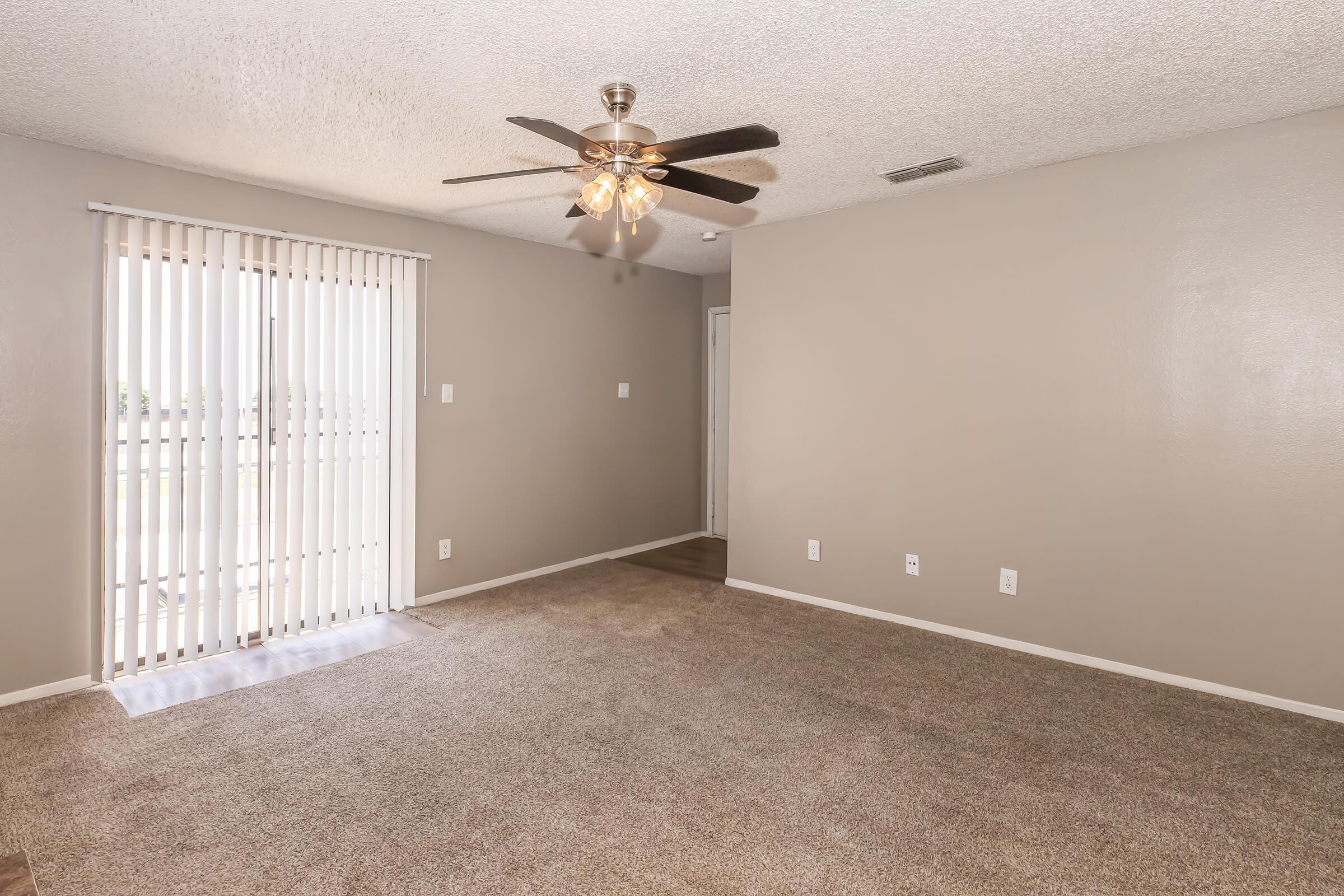 A spacious living room with beige walls and plush carpet, featuring a ceiling fan with light. There are vertical blinds on a large window, allowing natural light. A door leads to another room, creating a cozy and inviting atmosphere.