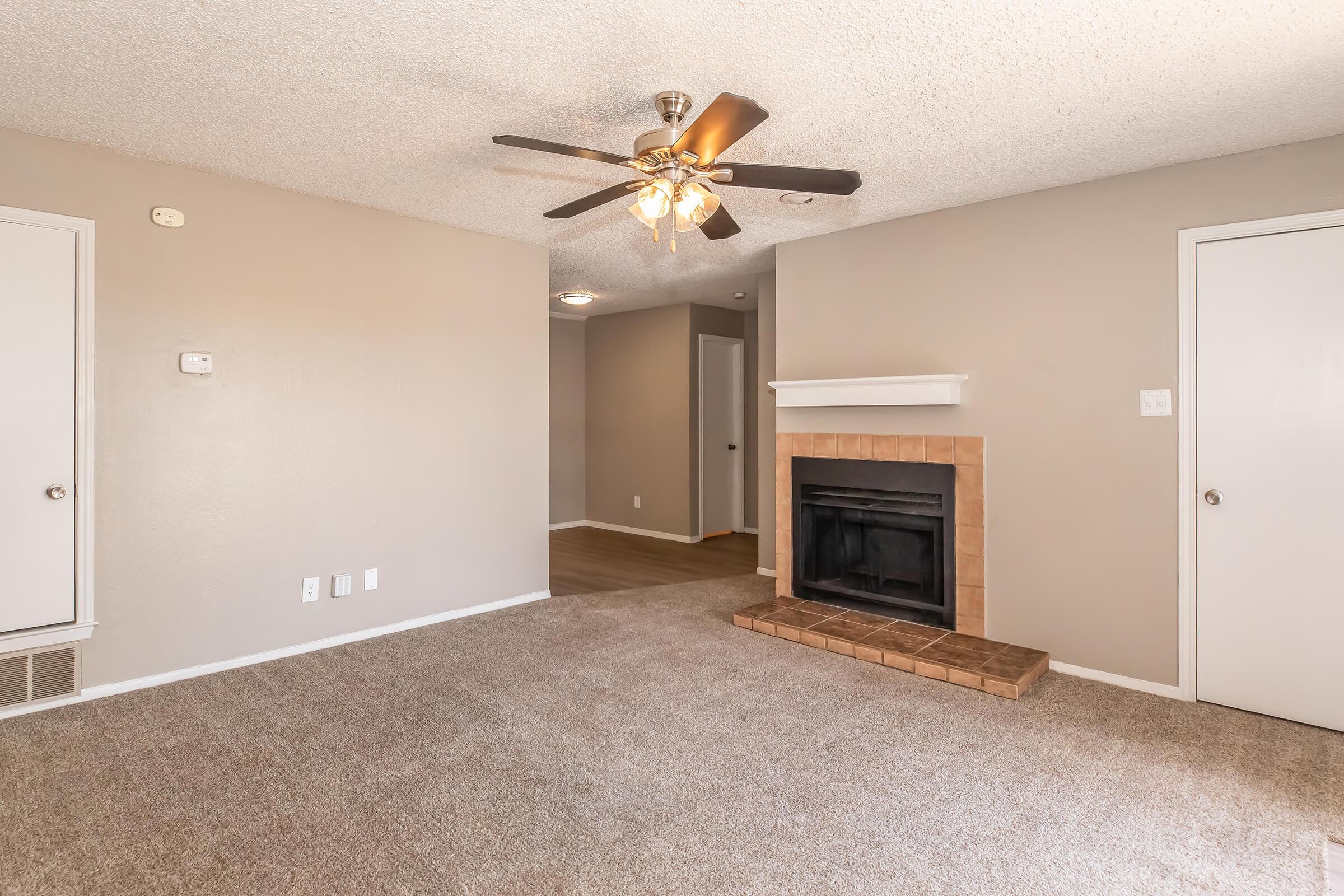A cozy living room featuring a ceiling fan, a decorative fireplace with a tiled hearth, and beige walls. The carpeted floor adds warmth, and there are white doors leading to other areas of the home. Natural light fills the space, creating an inviting atmosphere.