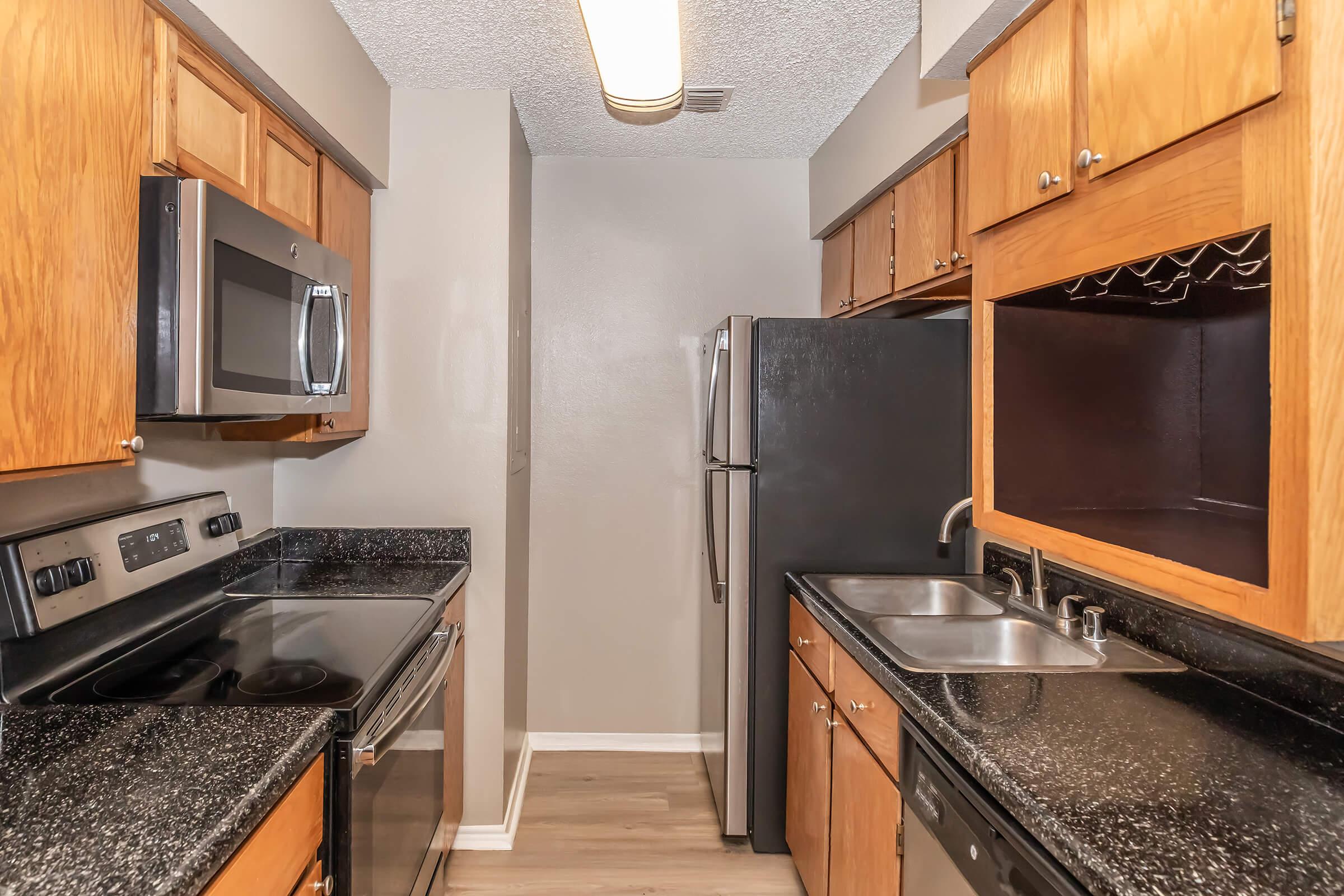 Modern kitchen with wood cabinets, stainless steel appliances including an oven, microwave, and refrigerator, black countertops, and a double sink. The walls are painted in a light color, and the flooring is a light wood laminate. There is ample cabinet storage and a well-lit space.