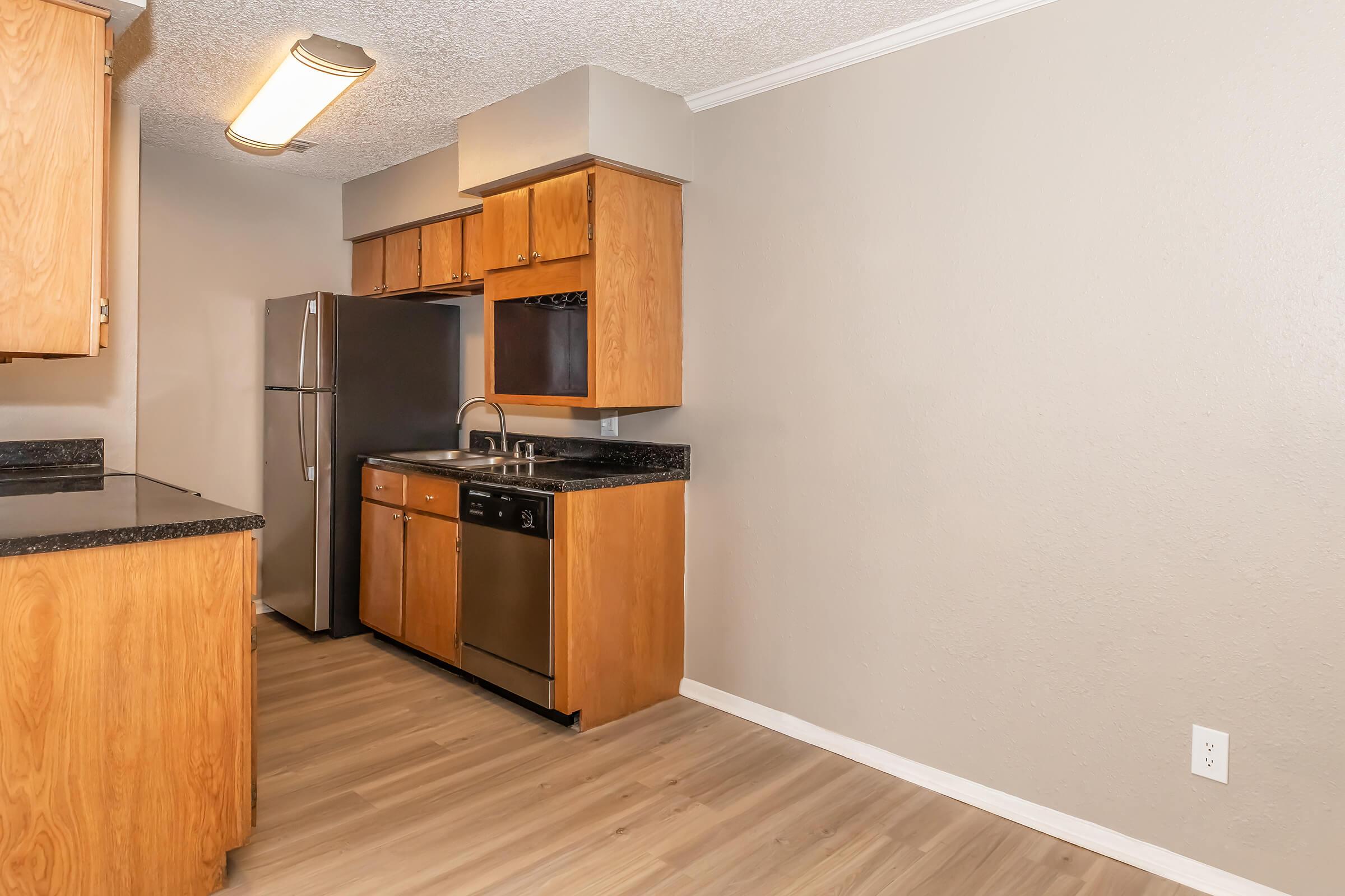 A modern kitchen featuring wooden cabinetry, a stainless steel refrigerator, a dishwasher, and a sink with a countertop. The flooring is light wood, and the walls are painted a neutral color. A ceiling light is visible, illuminating the space.