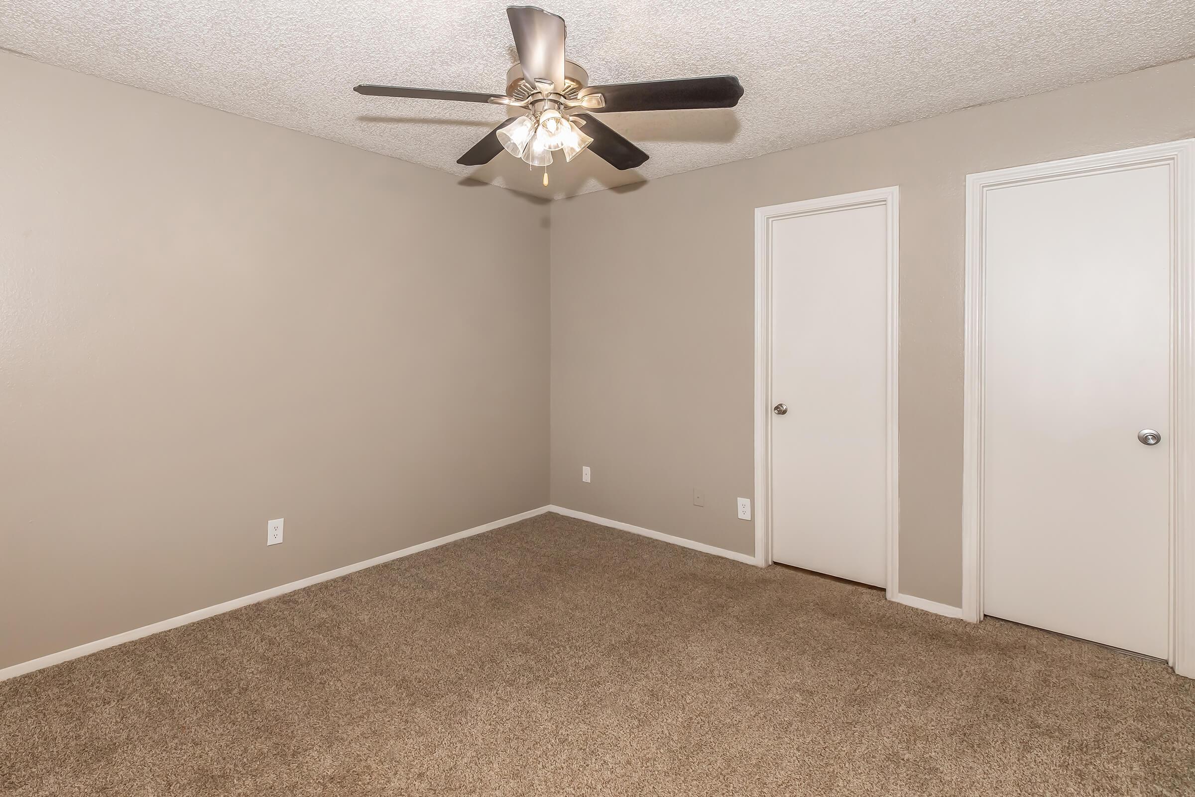 Empty room with light brown walls and plush beige carpet. A ceiling fan with five lights hangs in the center, and there are two white doors on the right side leading to closets. The room is well-lit and appears clean and ready for furnishing.