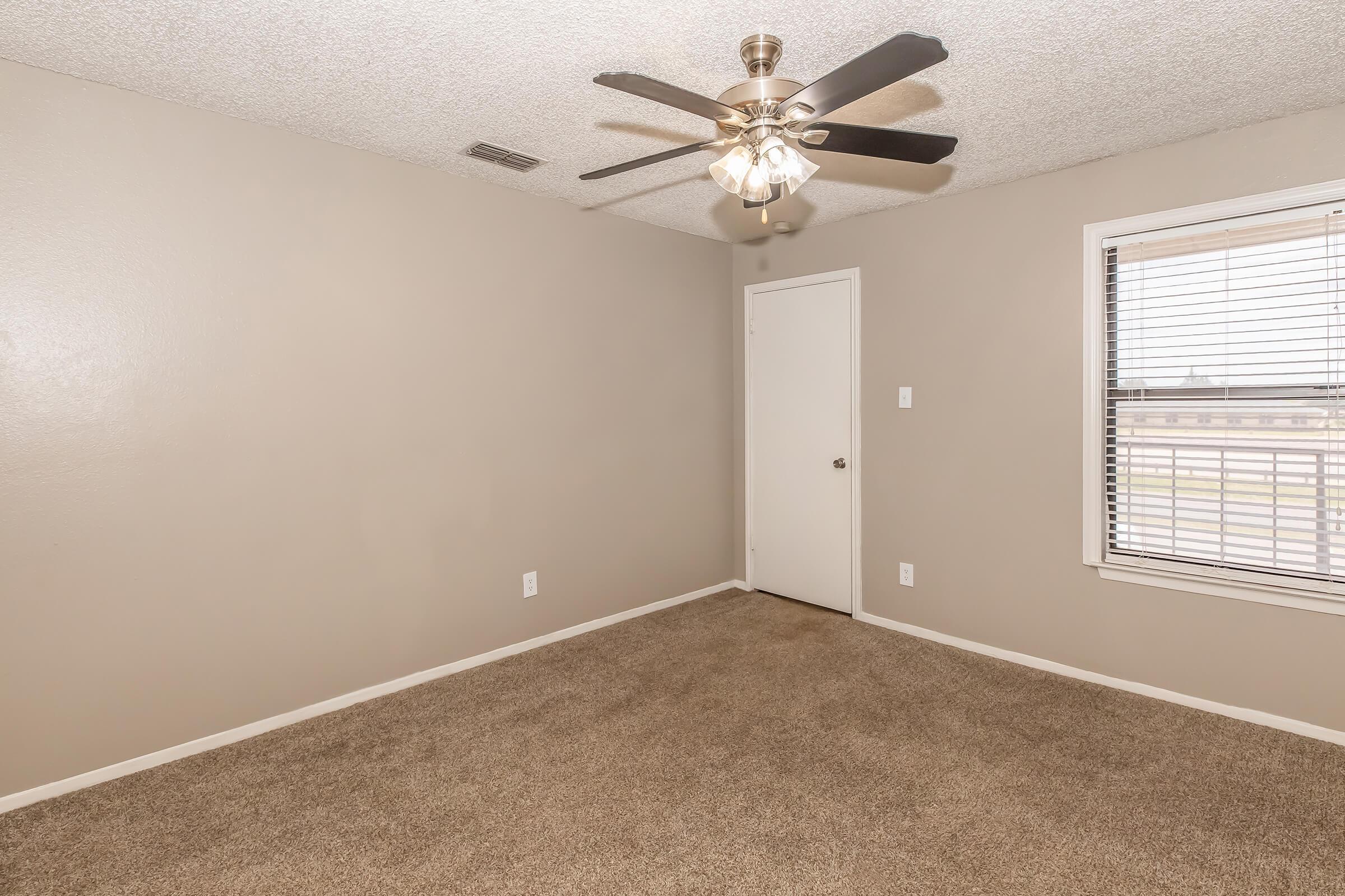 A small, empty bedroom featuring beige walls and carpet, a ceiling fan with lights, and a window with blinds. The room has a single door leading to another space, creating a minimalist and neutral atmosphere.