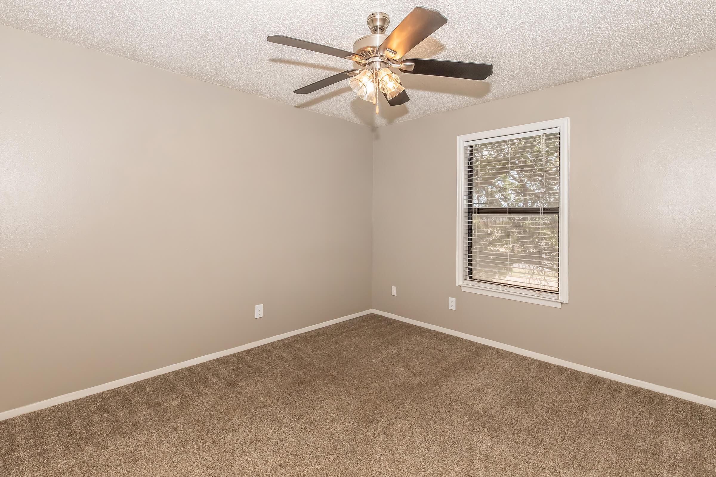 Empty room with light brown walls and a ceiling fan. The floor is covered in beige carpeting. A window with white blinds allows natural light in, offering a view of the outdoors.
