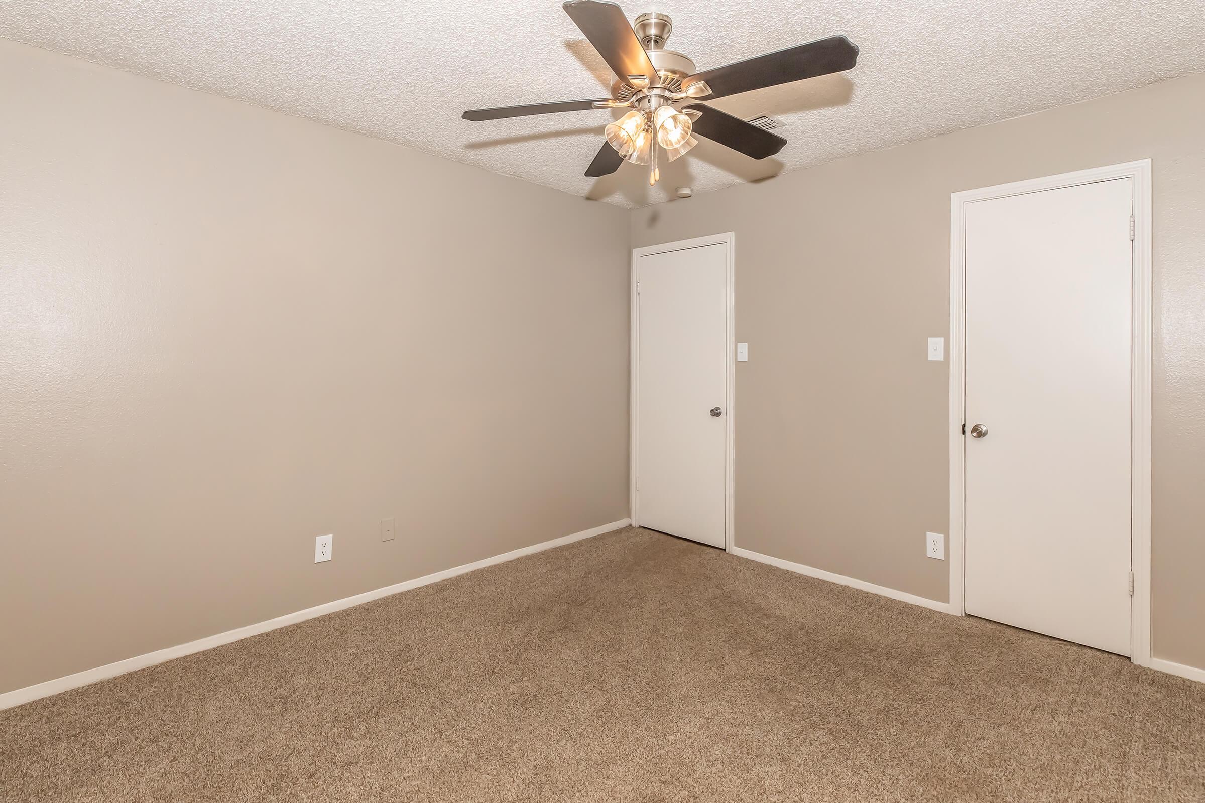 A spacious, empty room with light beige walls and carpeted flooring. It features a ceiling fan with lights and two doors—a closet and an entrance—on opposite walls. The room is well-lit and offers a clean, minimalist look.