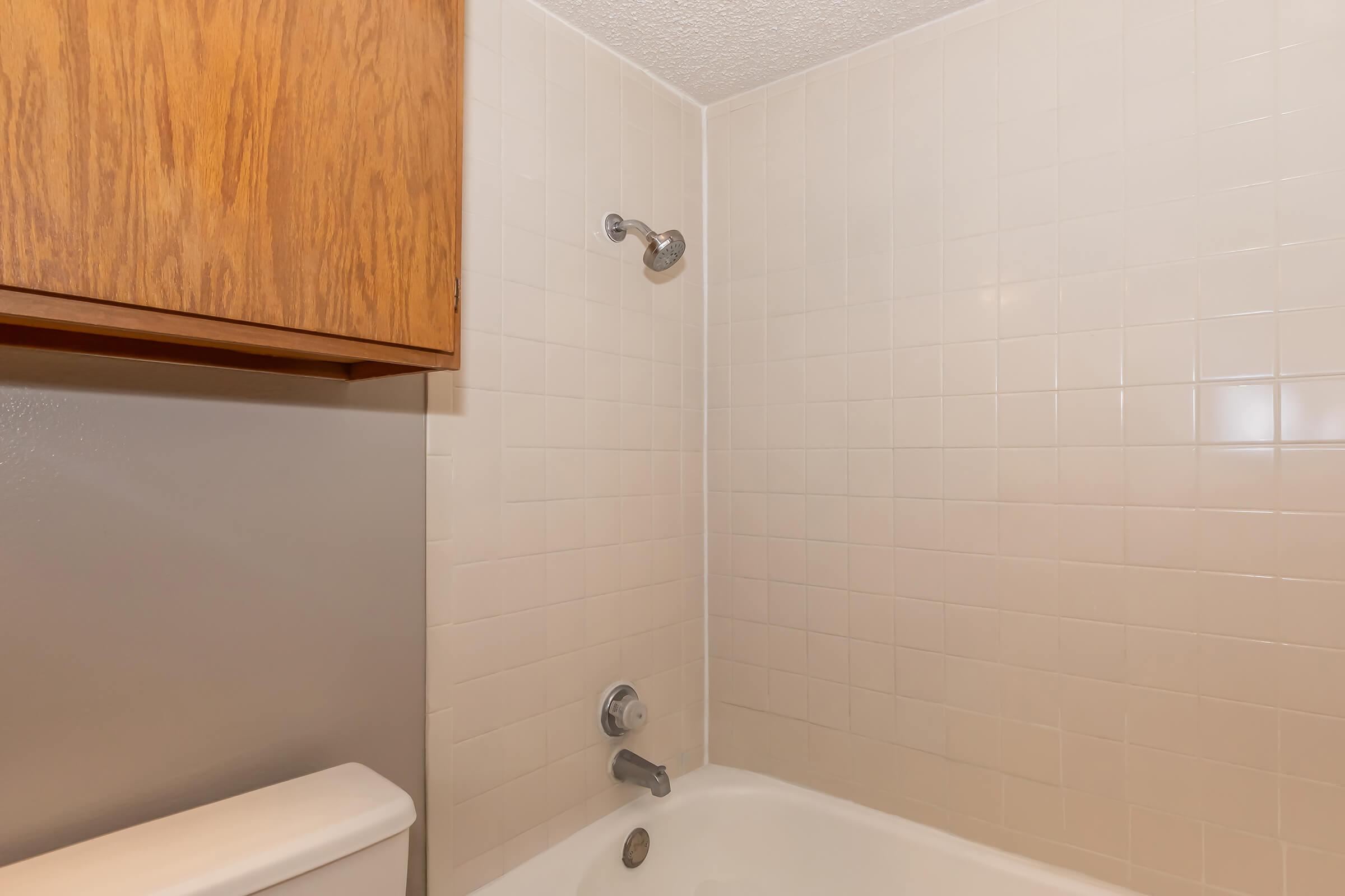 A clean and simple bathroom corner featuring a bathtub with a showerhead, surrounded by white tiled walls. There is a wooden cabinet above the tub and a toilet partially visible on the left side. The overall ambiance is bright and uncluttered.