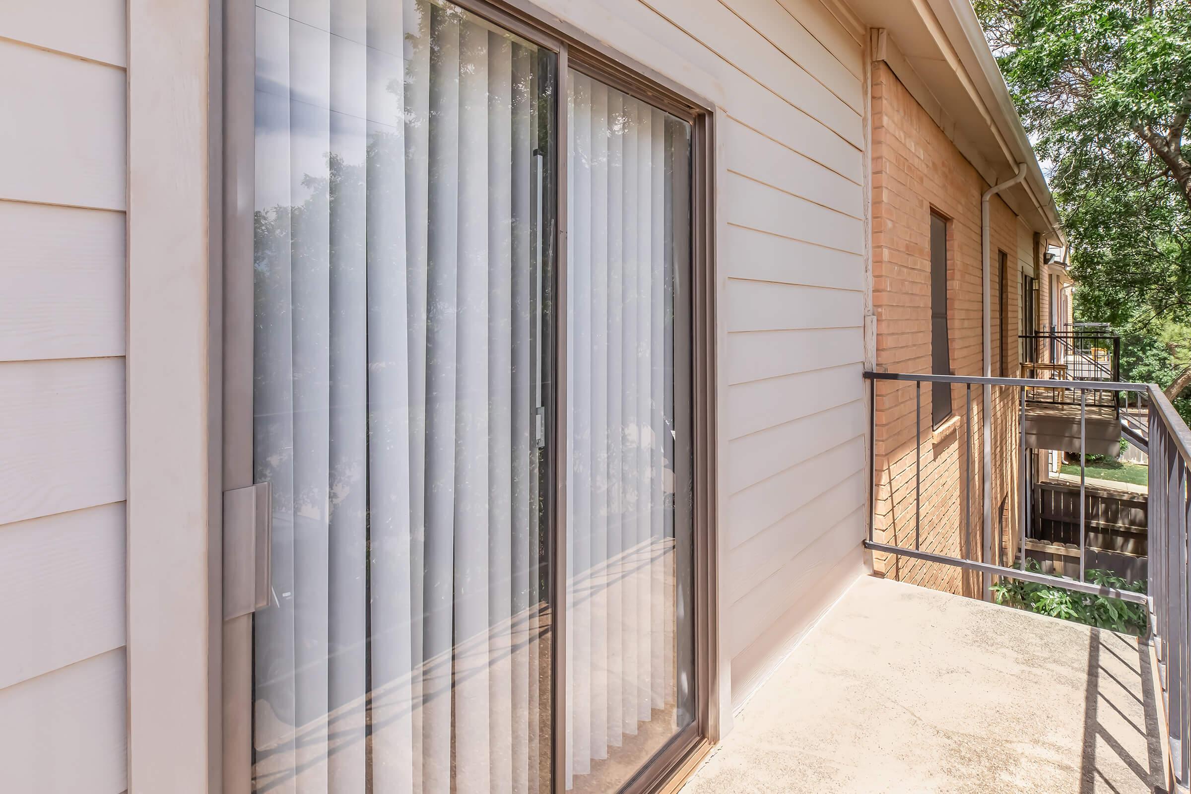 A view of a balcony with vertical blinds covering a sliding glass door. The exterior of the building features light-colored siding on one side and brick on the other. Lush greenery is visible beyond the railing, suggesting a natural outdoor setting.