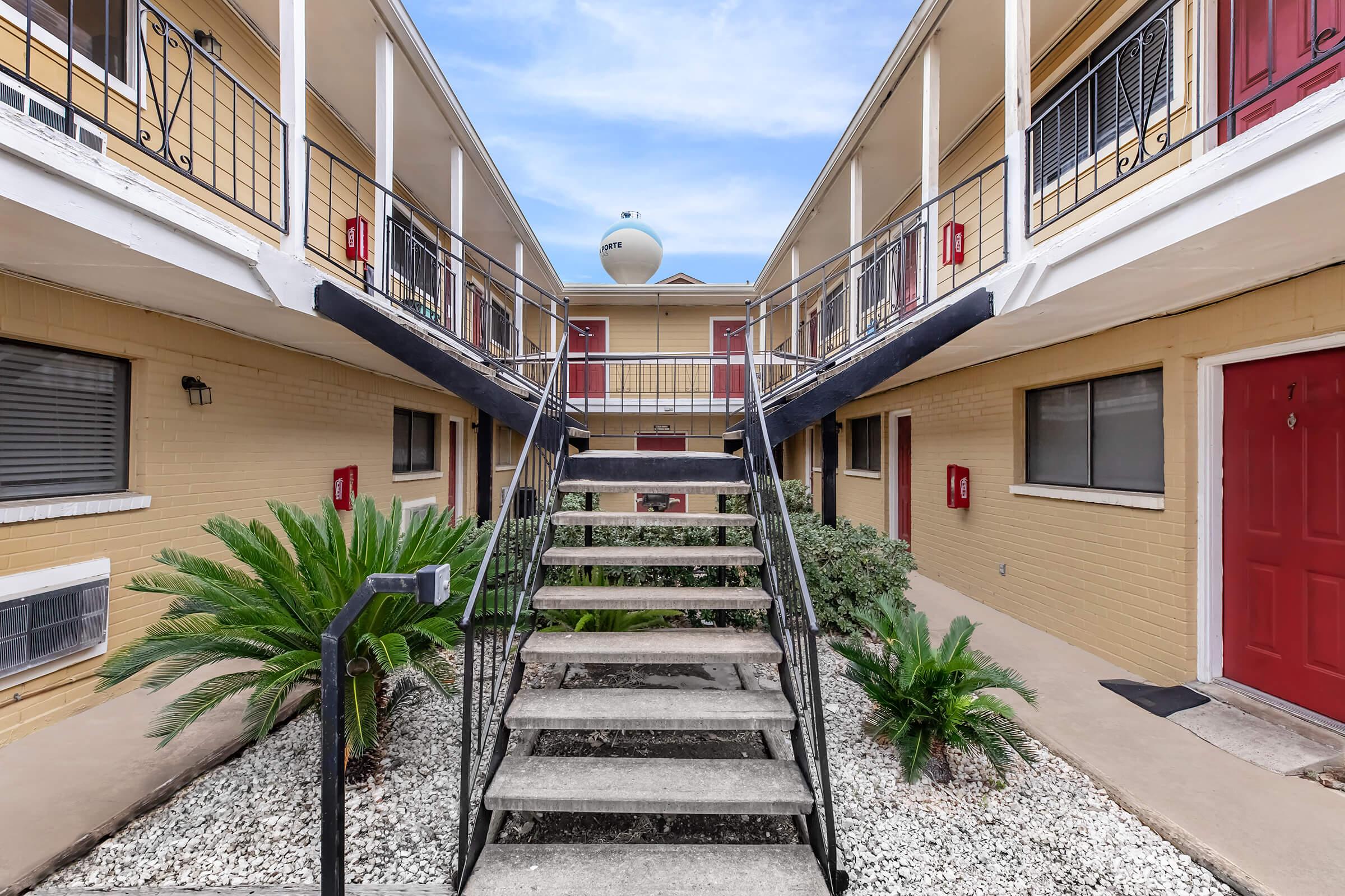 A view of an apartment complex courtyard featuring a central staircase leading to upper levels, surrounded by potted plants and gravel. There are several doors visible on the lower level, with a water tower visible in the background against a blue sky.