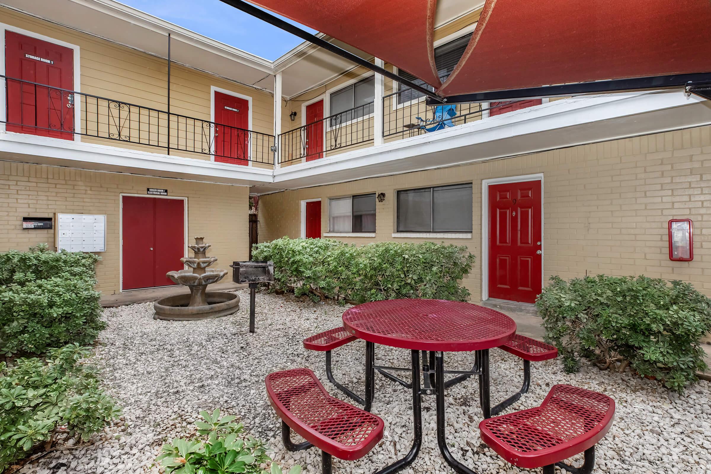 A courtyard with a red picnic table and benches surrounded by low shrubs, featuring a small fountain and a barbecue grill. The area is flanked by buildings with red doors on the upper and lower levels, providing a communal space in a residential setting.