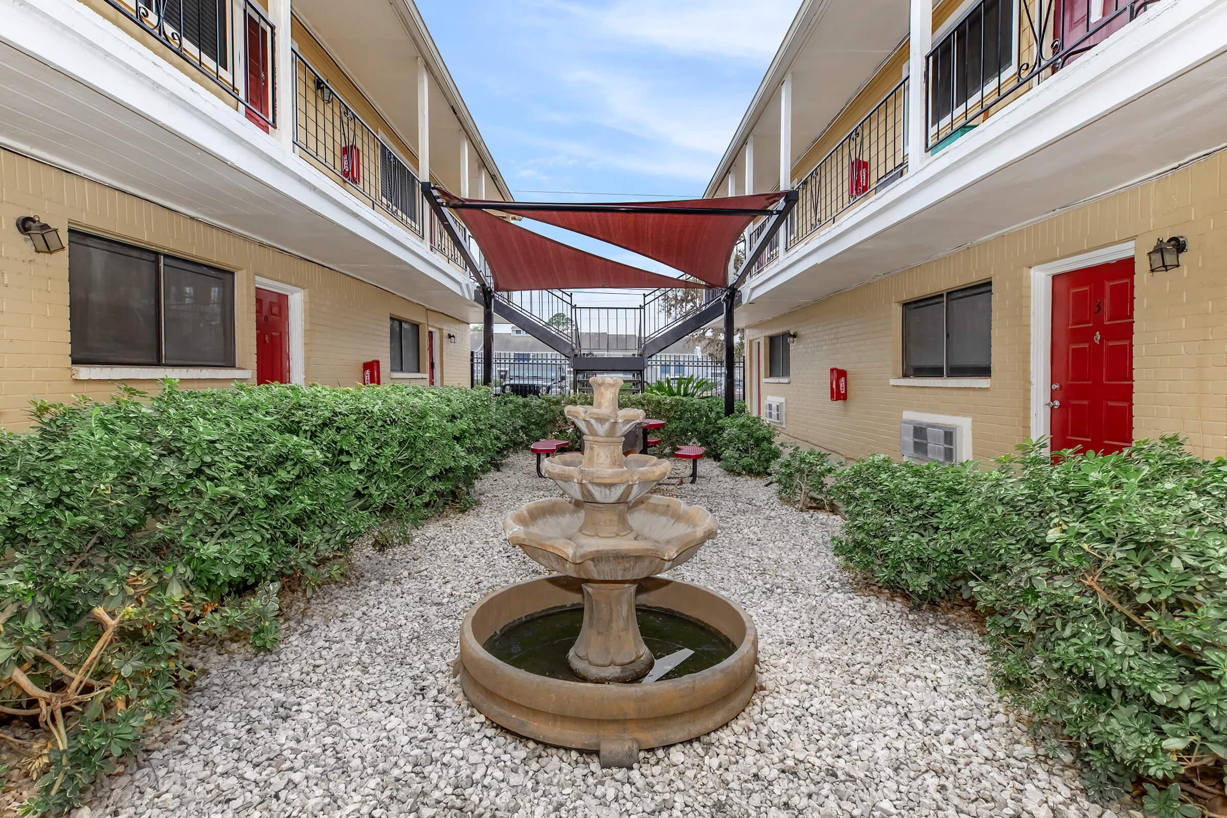 A courtyard view of an apartment complex featuring a central stone fountain surrounded by gravel and greenery. Balconies with red doors on either side overlook the area, and a shaded seating area is visible in the background under a red canopy.