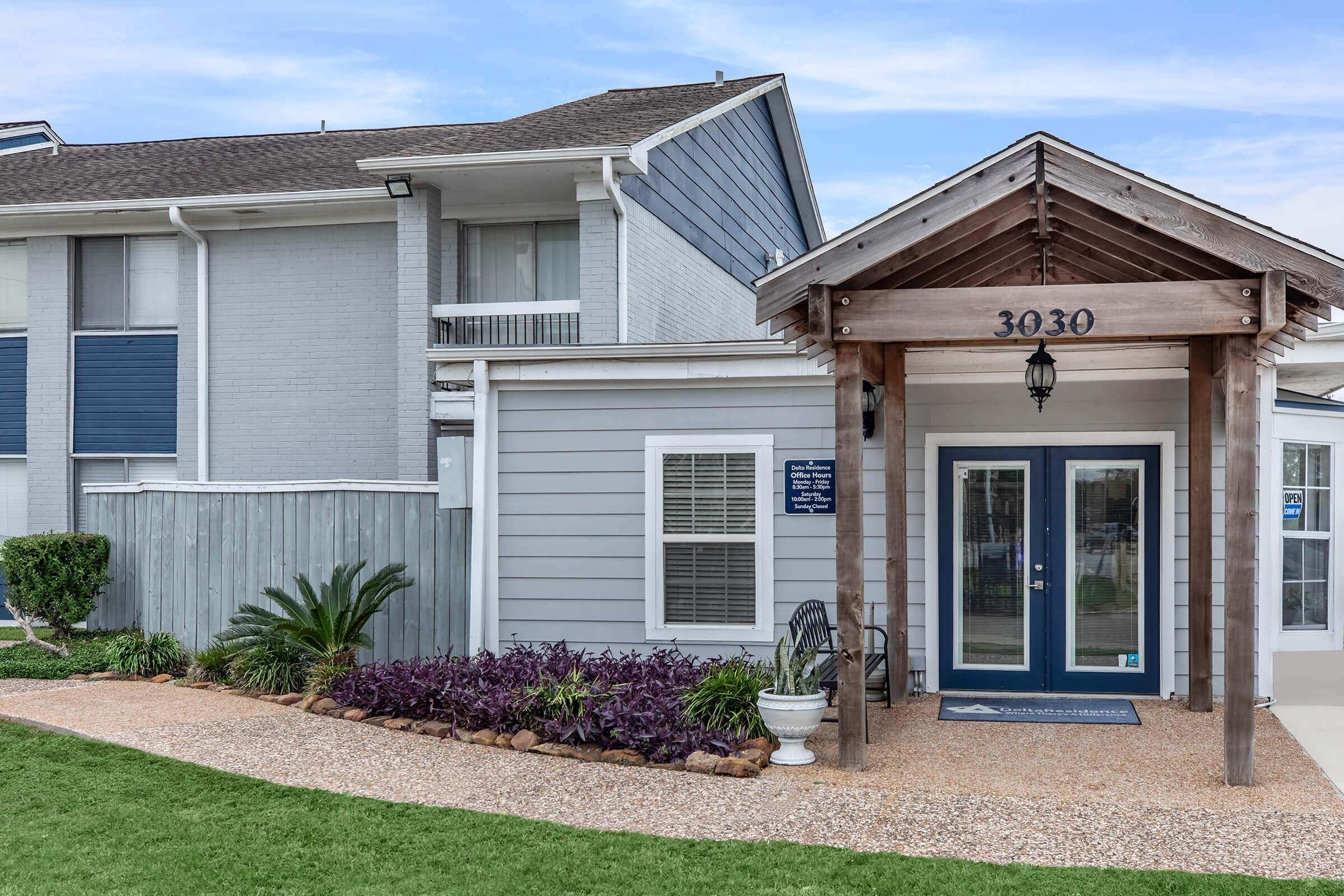 A welcoming entrance to a modern building featuring a wooden porch with a sign reading "3030." The facade has light blue and gray siding, large windows, and landscaped greenery. The pathway is lined with pebbles and plants, creating an inviting atmosphere.