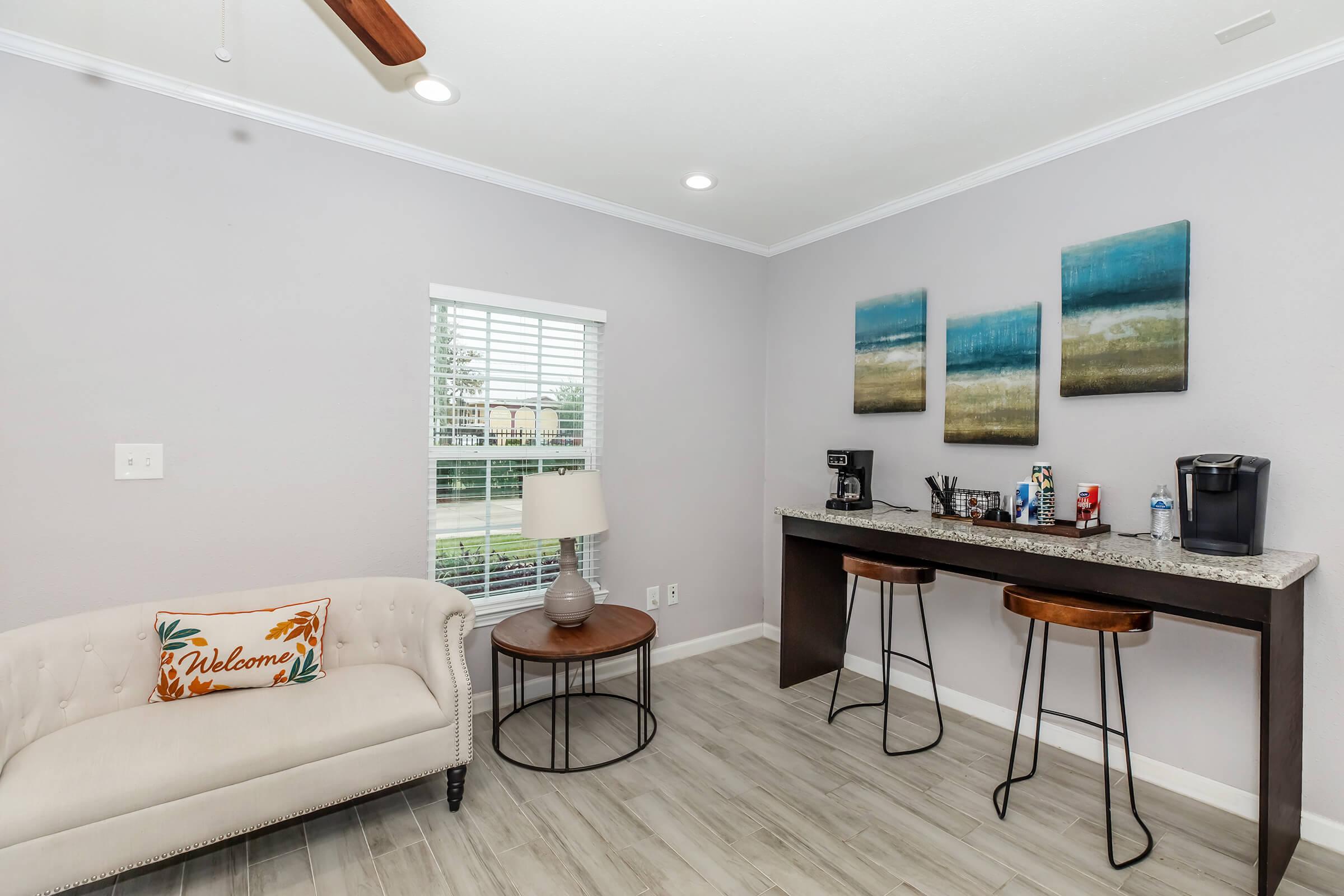 A cozy corner of a room featuring a light-colored sofa with a "Welcome" pillow, a round side table, a modern lamp, a bar-style counter with two stools, a coffee maker, and two bottles of water, along with three mounted paintings depicting tranquil blue and green landscapes. A window lets in natural light.