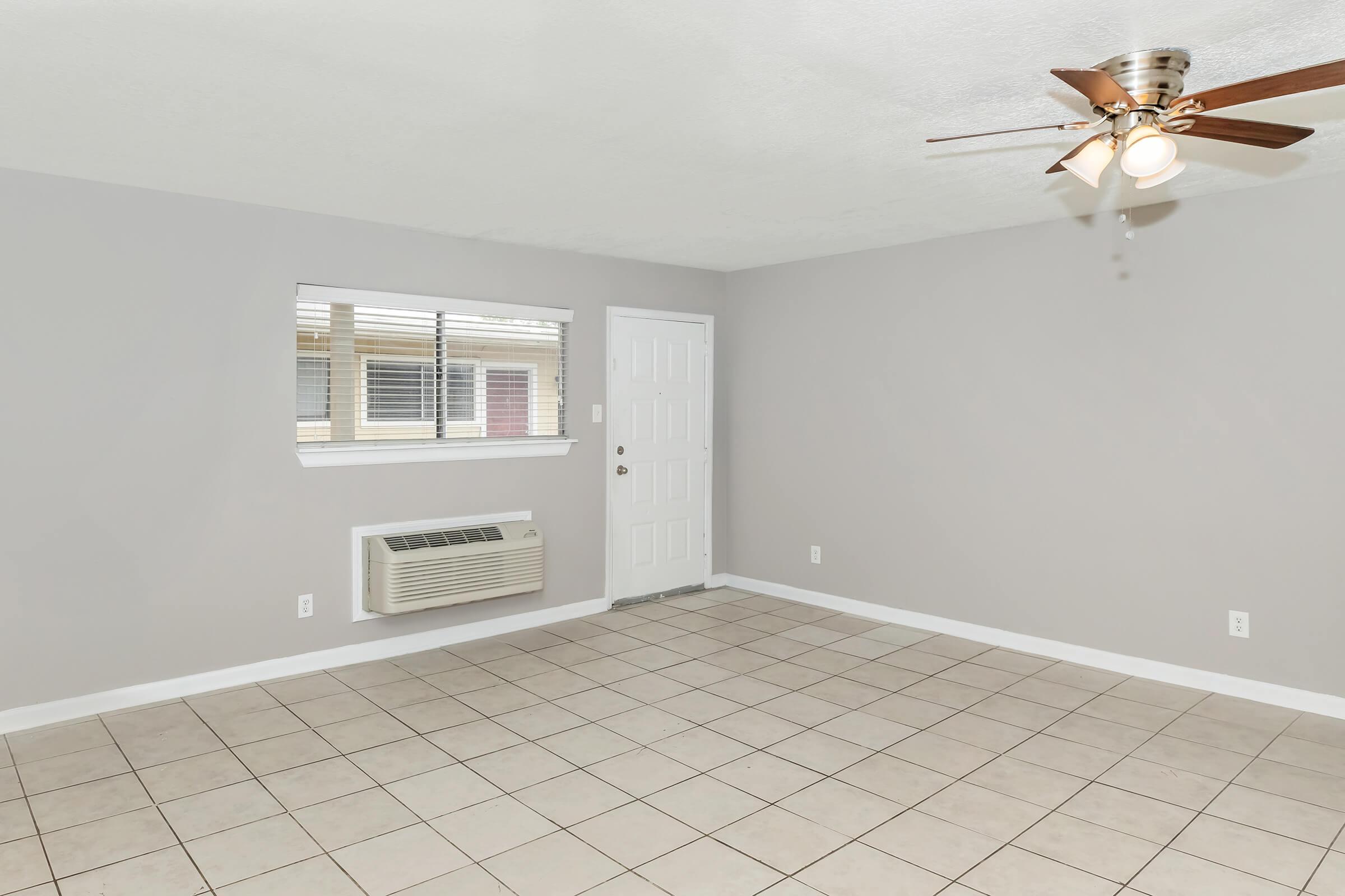 A spacious, empty living room with light gray walls and tiled flooring. It features a ceiling fan with light fixtures, an air conditioning unit on the wall, and a small window allowing natural light. The room has a white door leading outside, creating a bright and airy atmosphere.