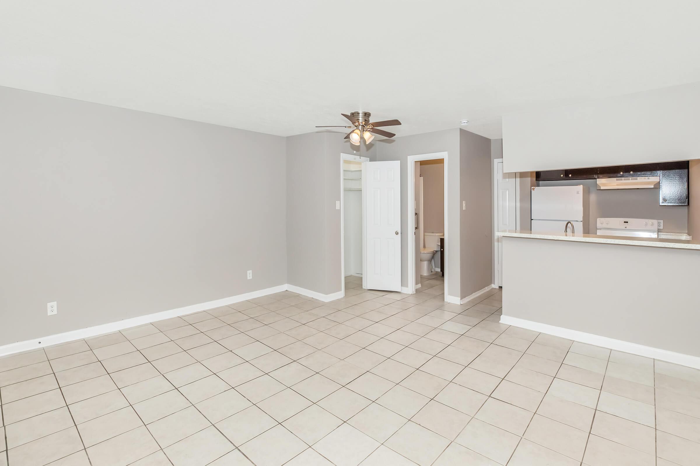 A spacious, light gray living area featuring tile flooring, a ceiling fan, and an open layout. A doorway leads to a small room with white walls, while the kitchen is partially visible in the background, showcasing a countertop and stainless-steel appliances.