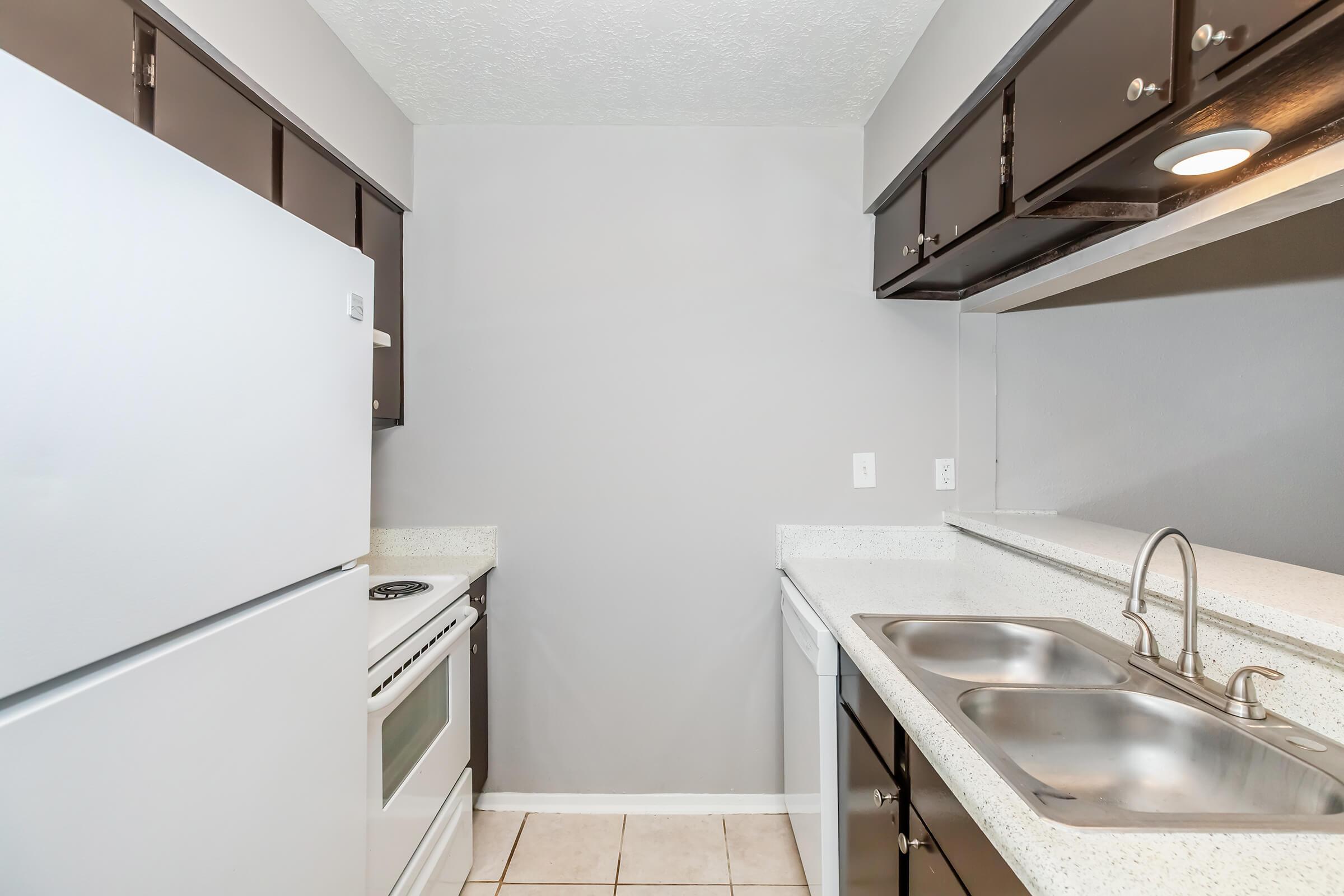 A modern kitchen featuring white appliances, including a refrigerator and oven, with dark cabinetry on the upper shelves. The countertops are light-colored, and there are two sinks. The walls are painted gray, providing a neutral backdrop to the space. The floor is tiled in light colors.