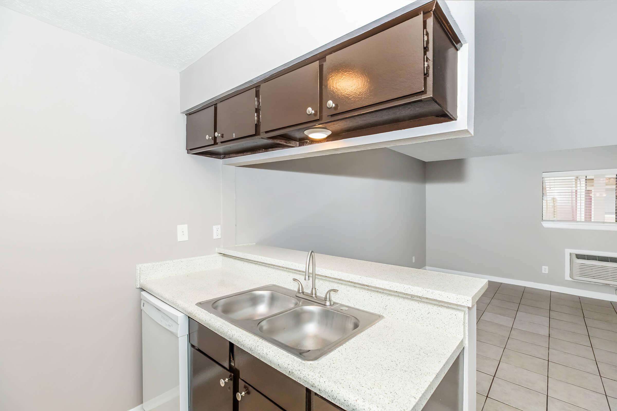 A modern kitchen featuring dark wooden cabinets above a light-colored countertop with a double sink. A dishwasher is visible to the left. The background shows a spacious living area with tile flooring and natural light coming through a window.