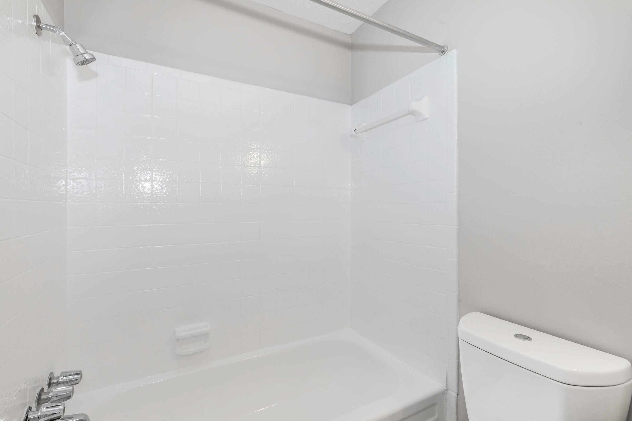 A clean, minimalistic bathroom with a white tiled shower area and a bathtub. The shower features a chrome showerhead and a chrome rail. To the right, there’s a modern white toilet, set against a plain, light gray wall. The overall appearance is bright and uncluttered.