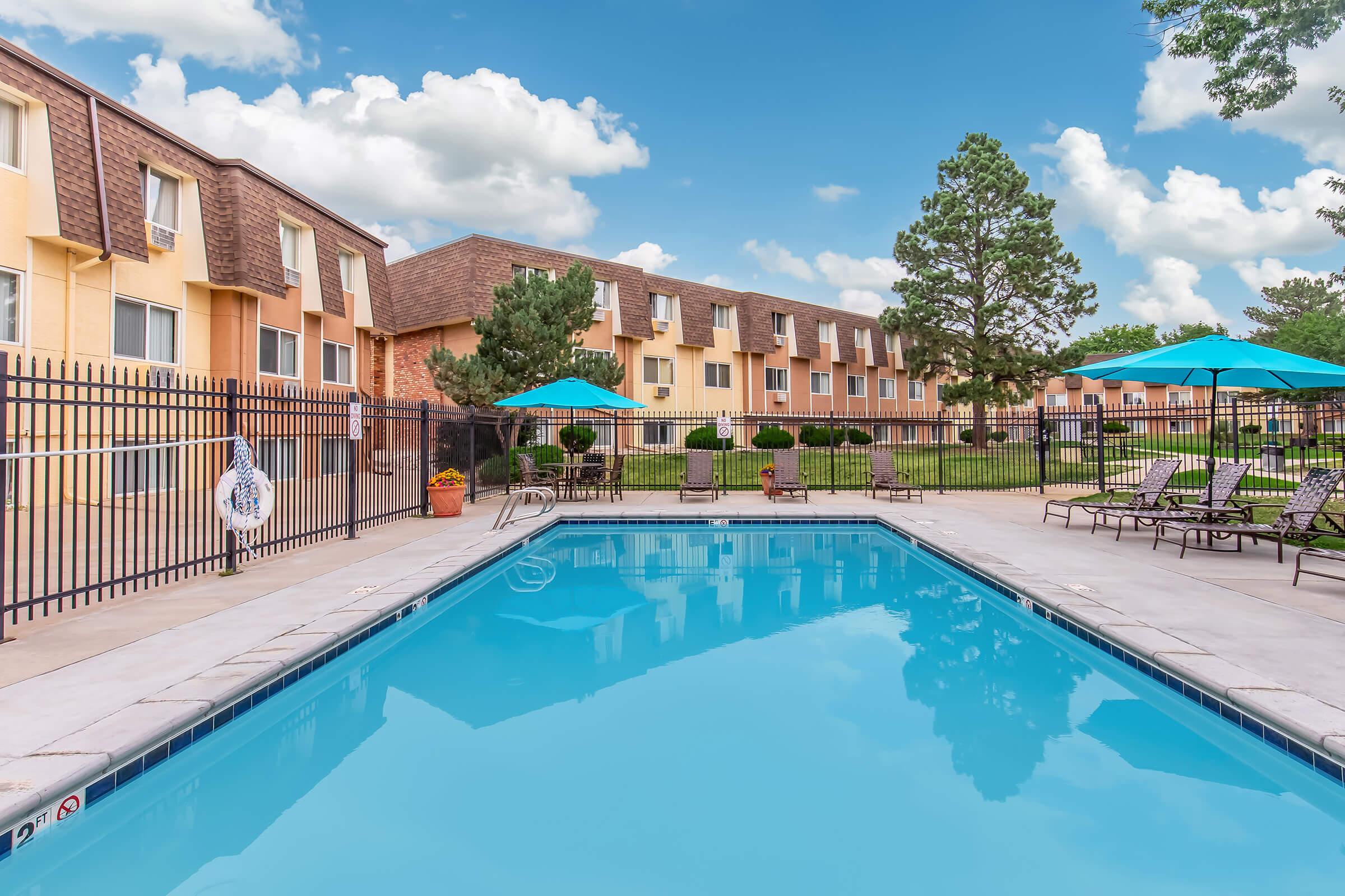 A clear blue swimming pool surrounded by lounge chairs and umbrellas, with a backdrop of a hotel building and green landscaping. Puffy white clouds are visible in a bright blue sky. The area is enclosed by a black fence, creating a welcoming outdoor space for relaxation.