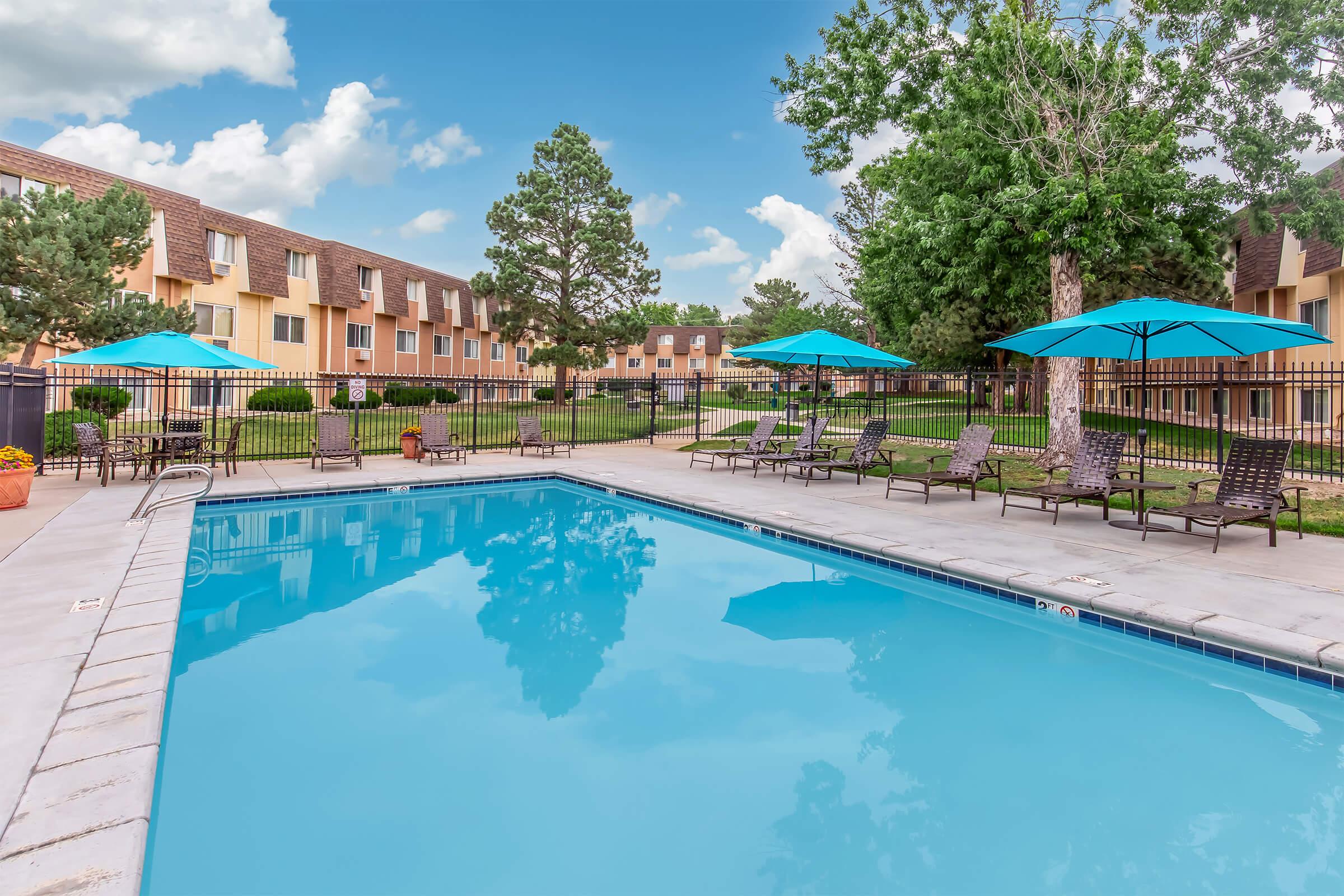 A swimming pool surrounded by lounge chairs and vibrant blue umbrellas. Lush green grass and trees can be seen in the background, along with a row of tan buildings. The sky is partly cloudy, creating a serene and inviting atmosphere. Ideal for relaxation and leisure activities.
