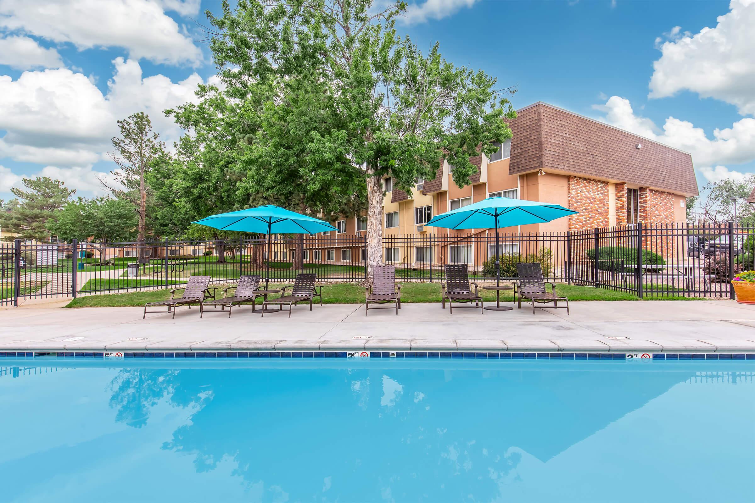 Outdoor swimming pool with a clear blue surface, surrounded by lounge chairs and large umbrellas. In the background, there is a brick building with multiple windows and green trees. The scene is set under a bright blue sky with fluffy white clouds.