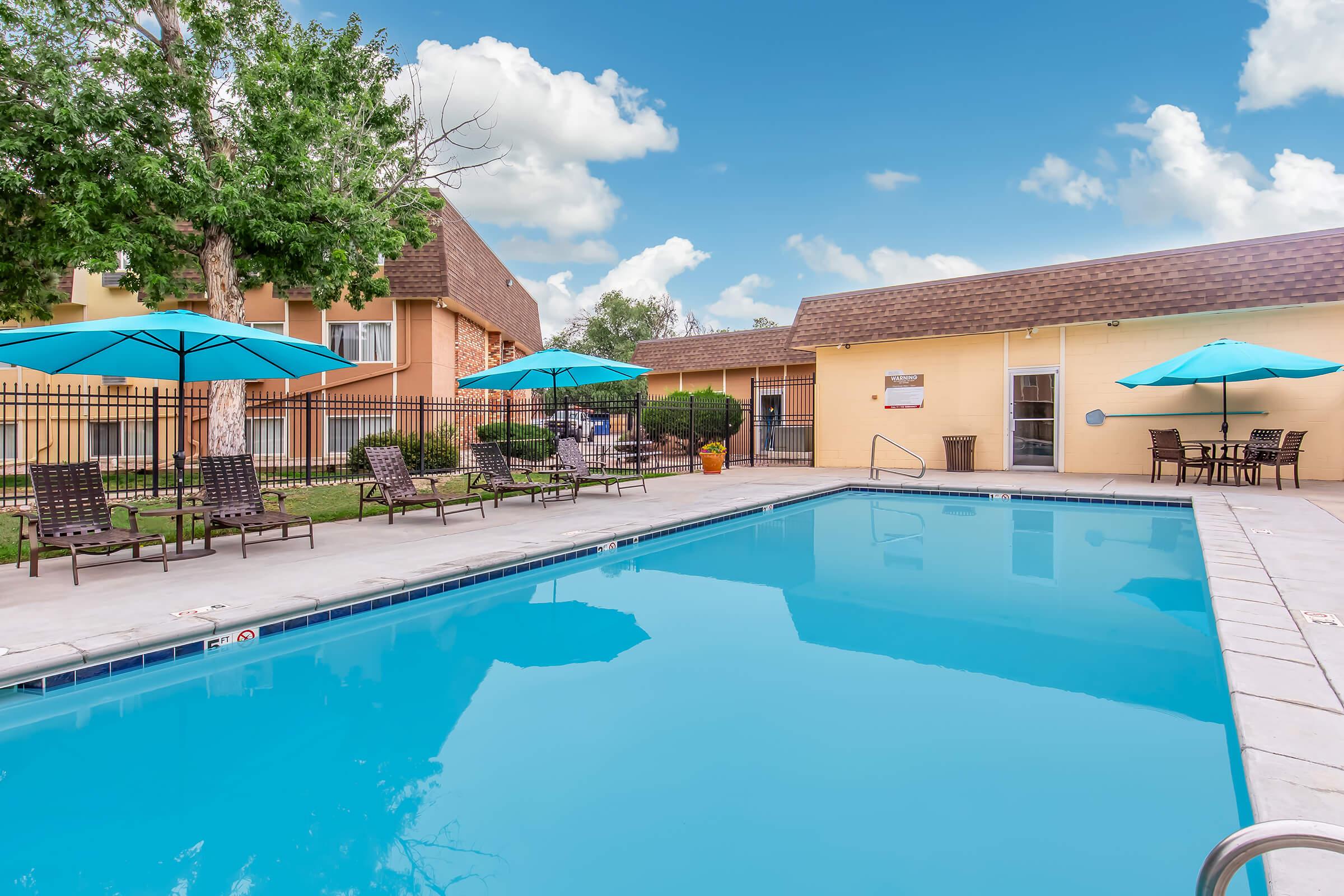 A clear blue swimming pool surrounded by lounge chairs and turquoise umbrellas. Lush trees are in the background, and there is a nearby building with a brown roof. The scene is set against a bright blue sky with fluffy white clouds.