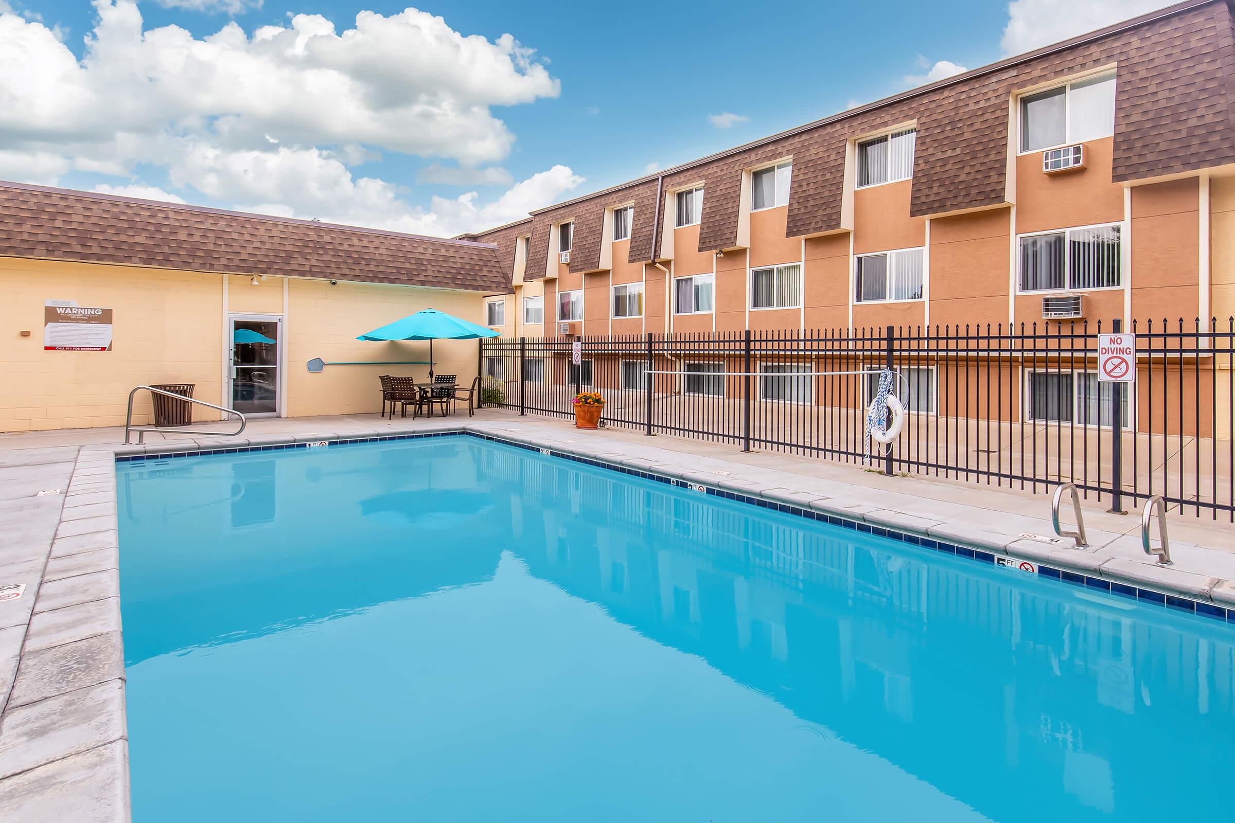 A clear swimming pool in the foreground with a bright blue surface, surrounded by a tiled deck. In the background, a three-story building with orange walls and brown shingled roofs. An umbrella-covered table is nearby, along with flowers and a lifebuoy on the fence. The sky is partly cloudy, enhancing the cheerful atmosphere.