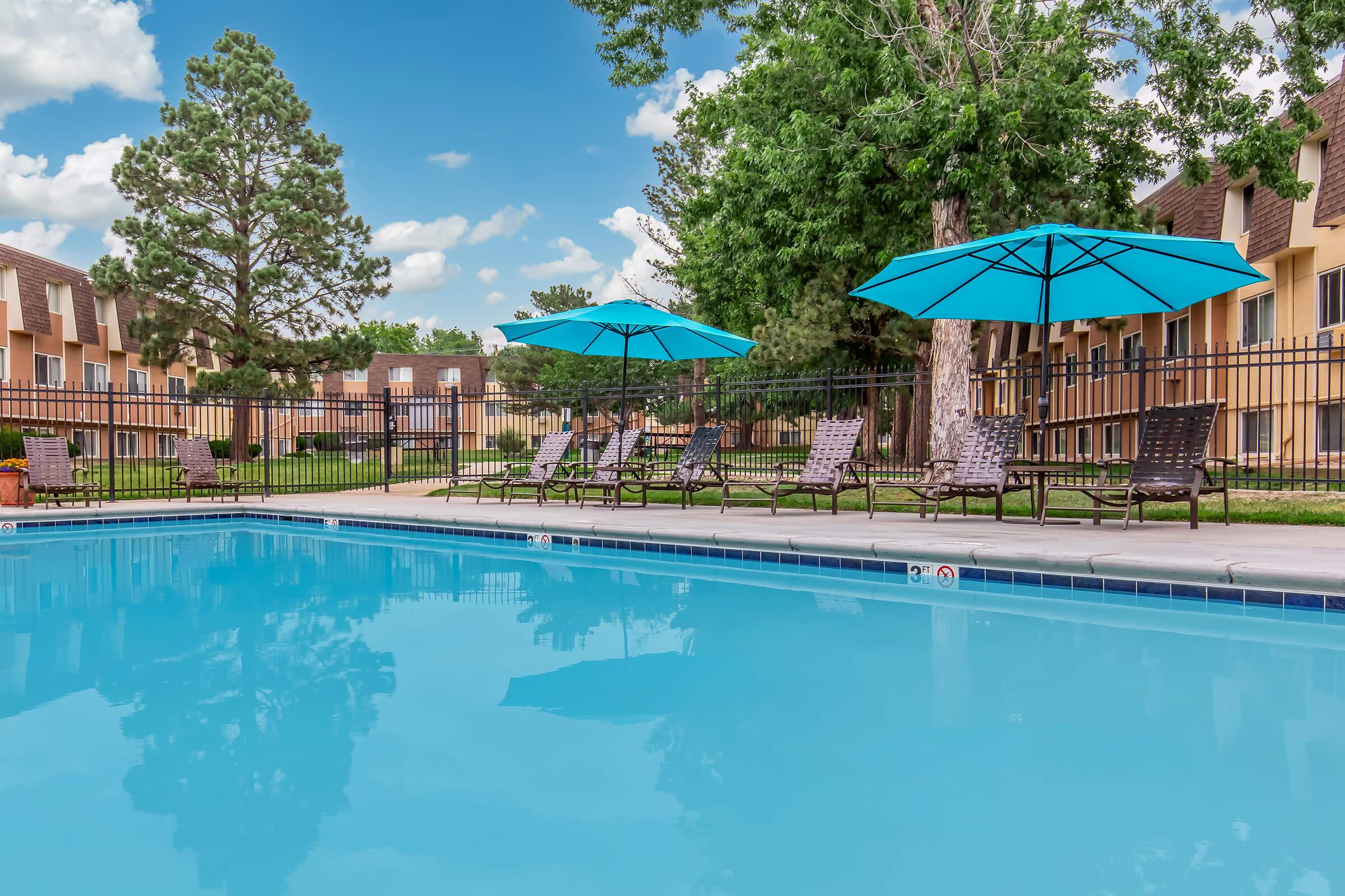 A serene swimming pool surrounded by lounge chairs and large blue umbrellas, set within a landscaped area. Grass and trees provide shade in the background, while the sky is partly cloudy, creating a relaxing atmosphere ideal for leisure and enjoyment.